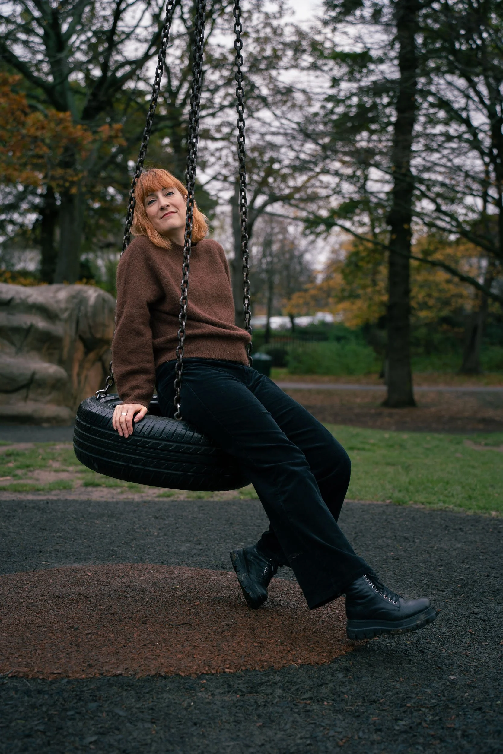 A woman with red hair in a brown sweater and black pants sitting on a tire swing in a park during autumn, with trees in the background.