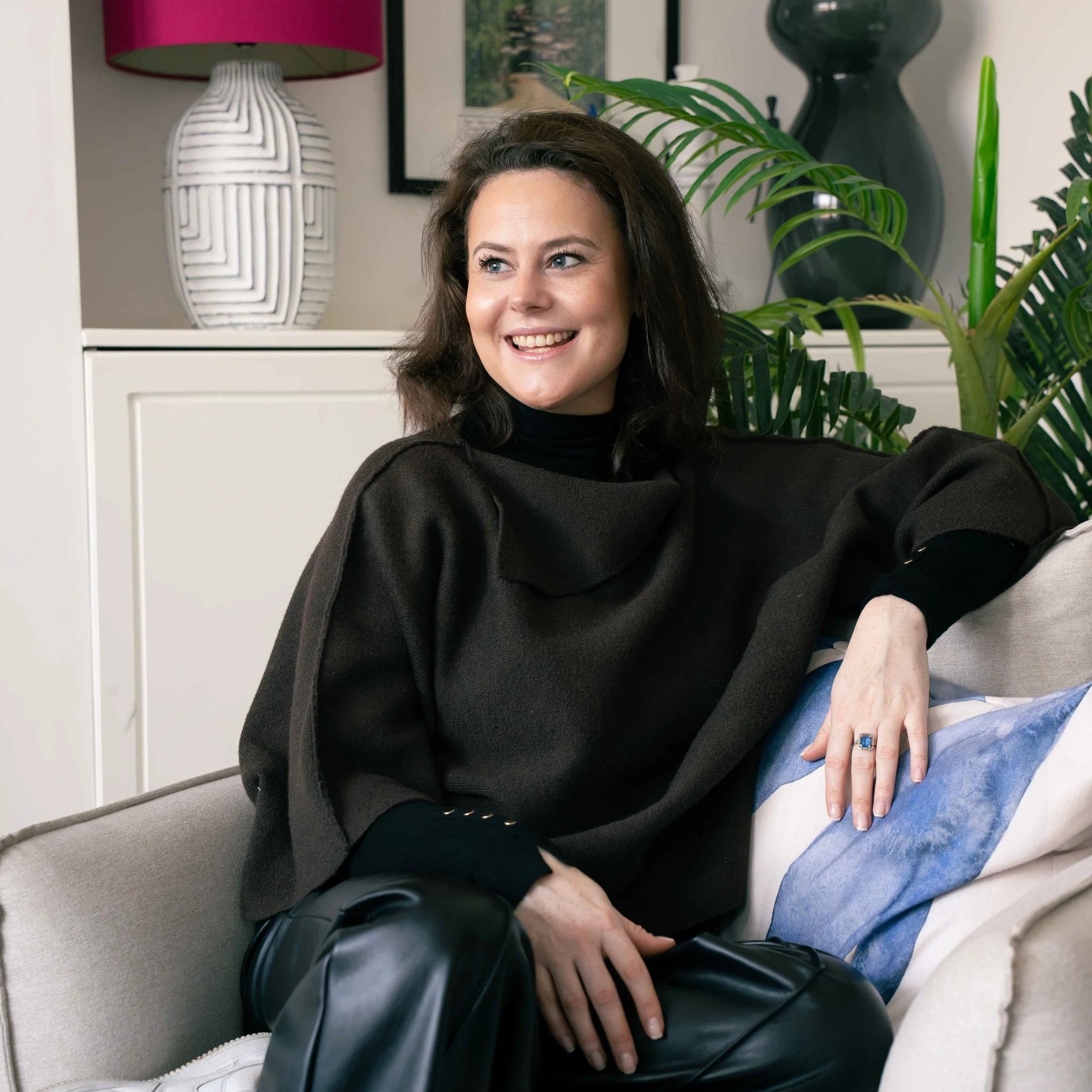 A woman with dark brown hair, wearing a black shirt, smiling and sitting on a couch next to a man in a beige hoodie. They are in a living room with a large green leafy plant behind them, a white cabinet, and decorative vases and lamps.