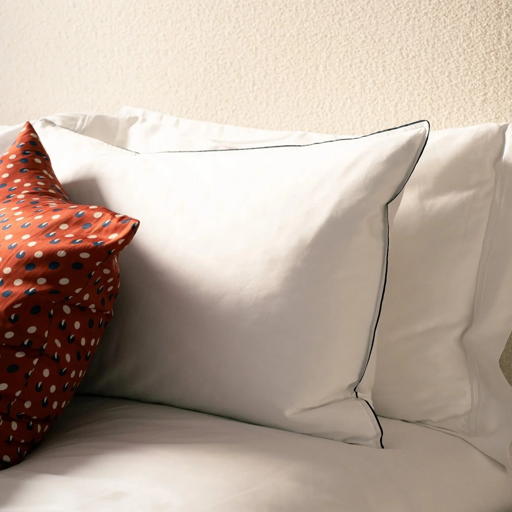 Close-up of a bed with white pillows embroidered with black piping and a red decorative pillow with a polka dot pattern