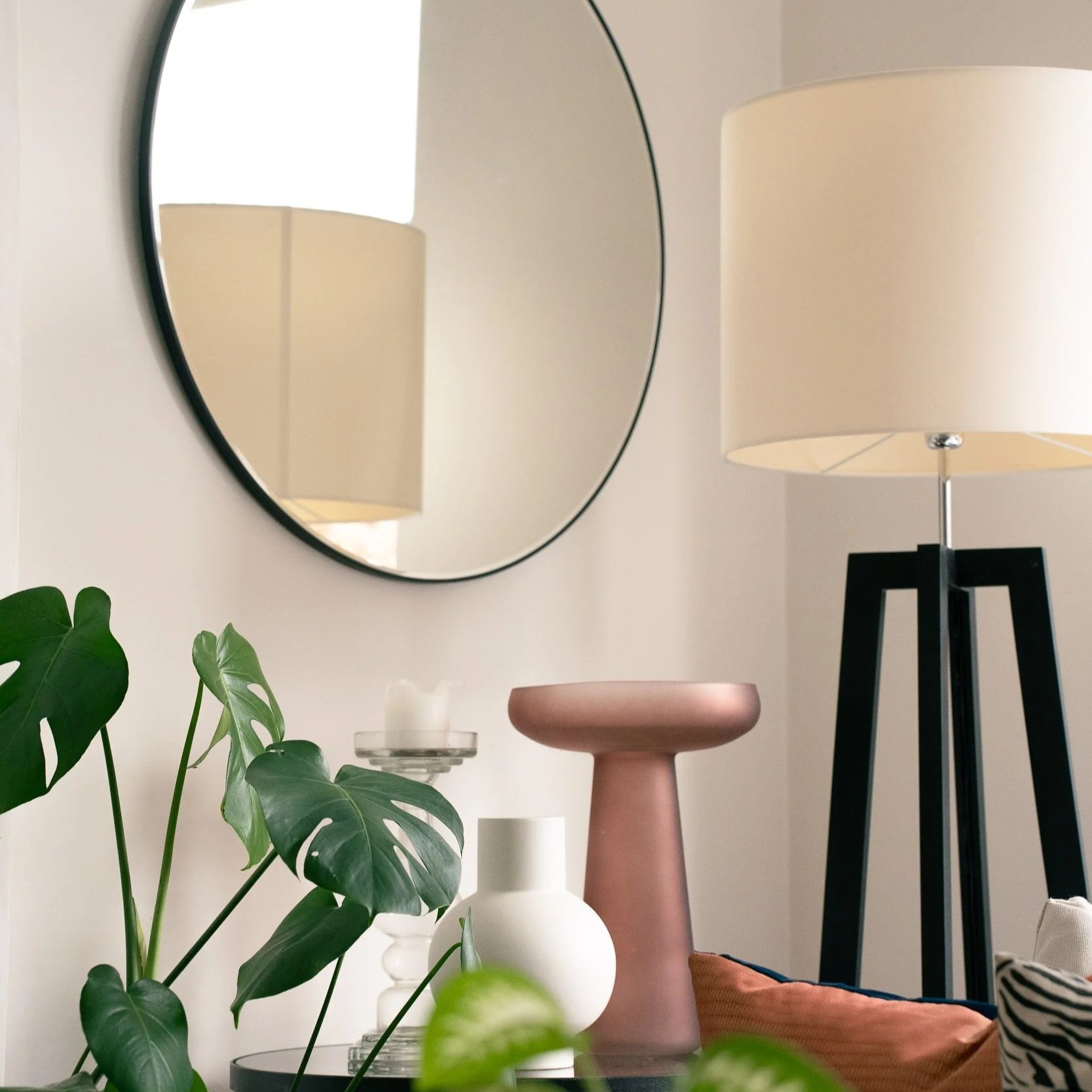 Decorative interior scene with a round mirror on the wall reflecting a beige lampshade, a black lamp stand, and a small viewing platform with vases, a pink ceramic vase, and a green plant with large leaves on a white surface.