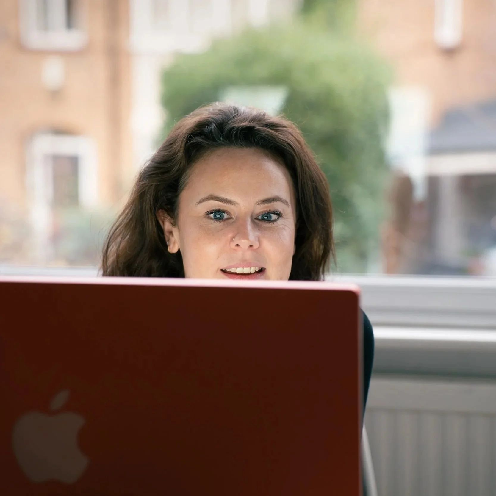 A woman with dark brown hair and blue eyes looking at a laptop in a bright room with large windows and blurred background of buildings and greenery outside.