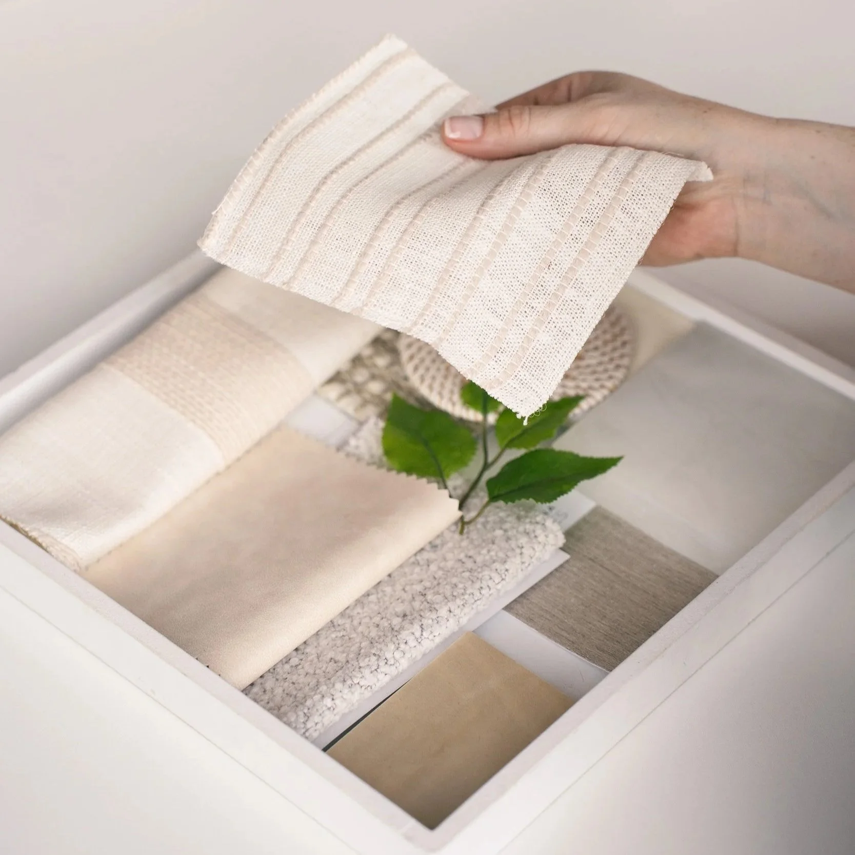 A white box containing various textured fabric samples, a small plant, and decorative stones.