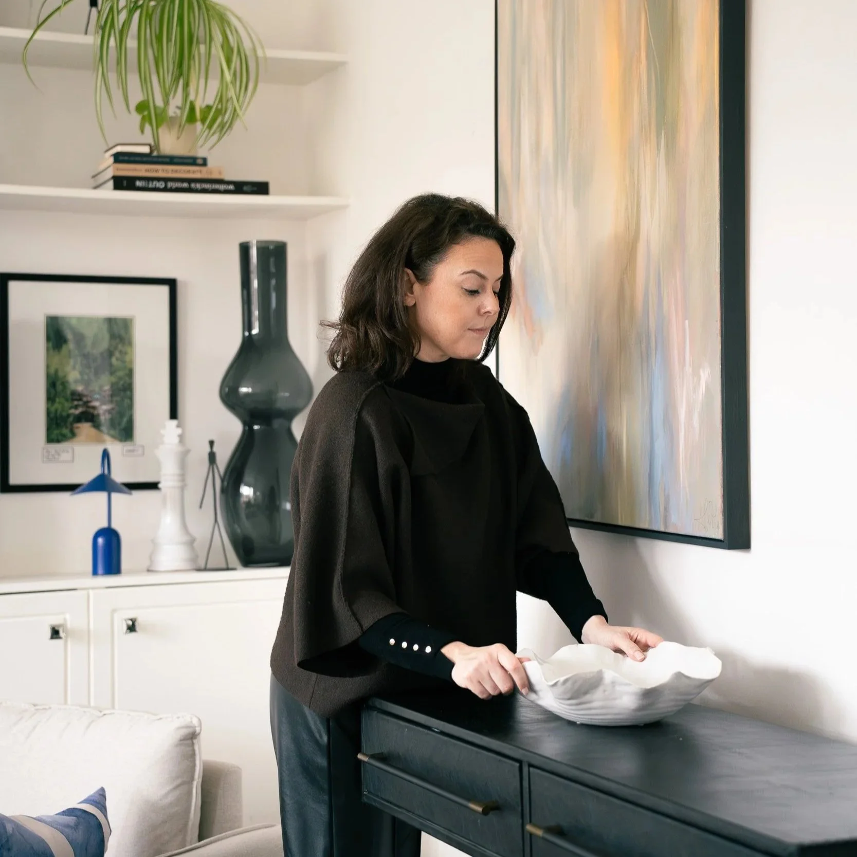 A woman with dark hair wearing a black top with silver buttons on the sleeves, standing at a dark-colored table, holding a white object in a decorative white bowl, in a modern room with artwork and plants.