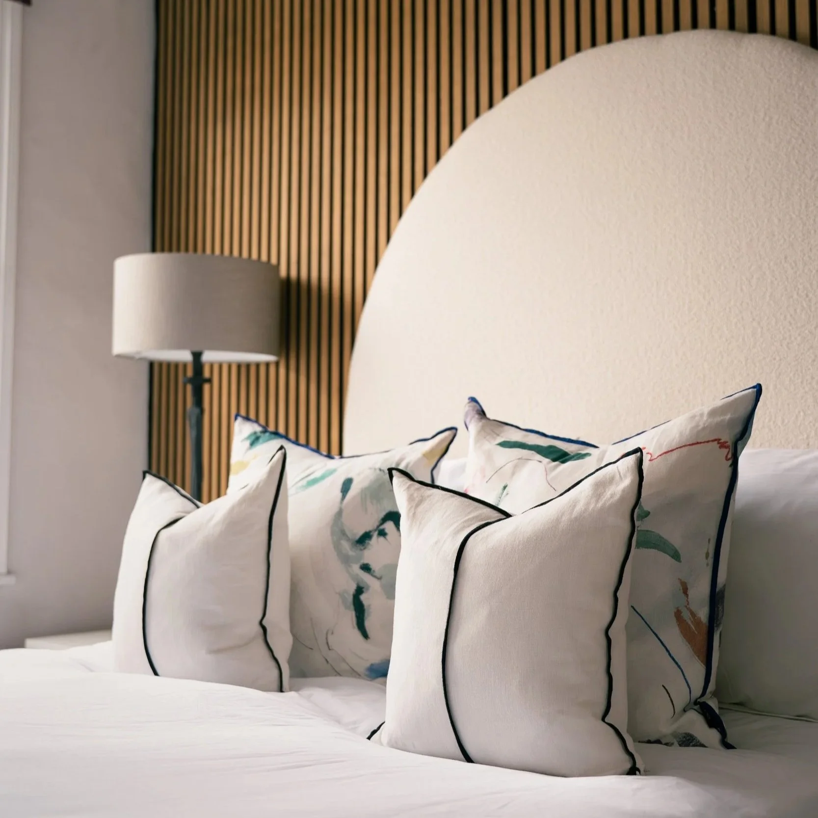 A bedroom with a bed featuring four decorative throw pillows with colorful, abstract patterns and black piping, set against a curved, beige headboard and a wooden slat wall, with a white table lamp on the bedside.