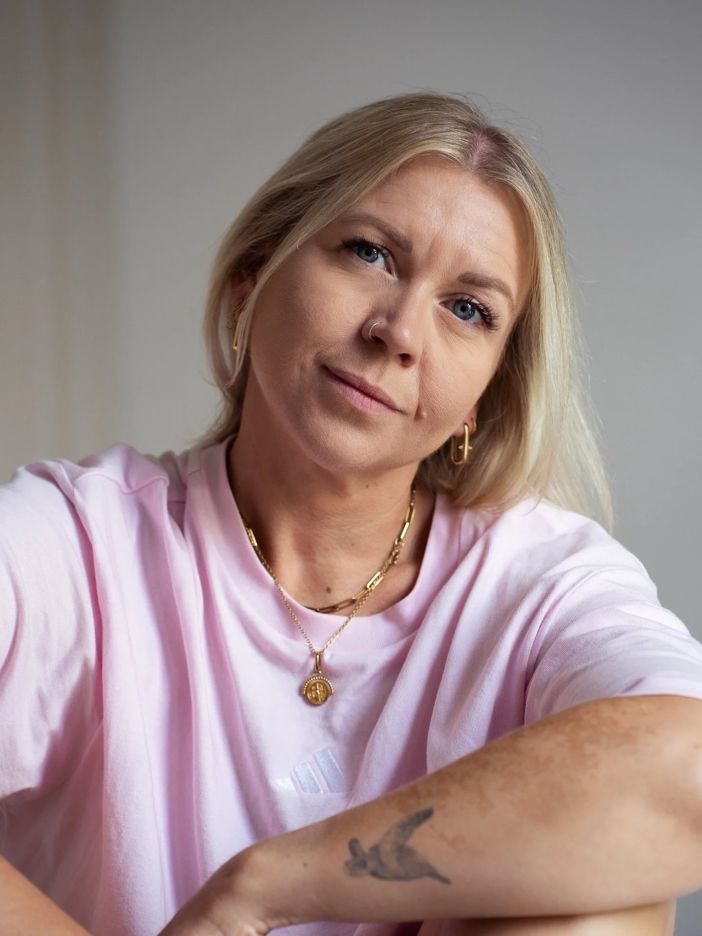 A woman with blonde hair and blue eyes taking a selfie. She is wearing gold jewelry, including hoops, layered necklaces, and a pendant. She has a nose ring and a tattoo on her arm. She is dressed in a light pink shirt.