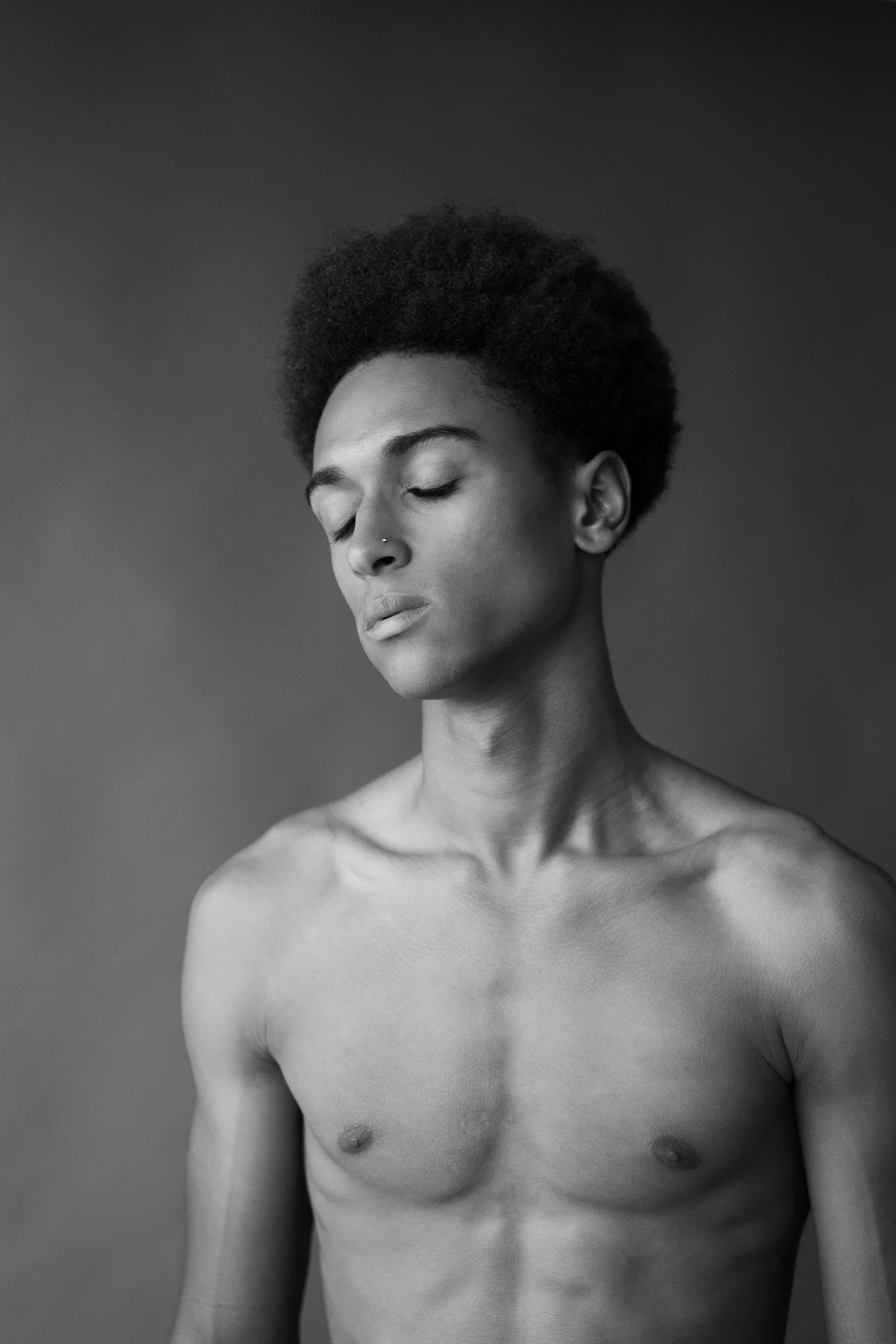 Black and white portrait of a topless woman with eyes closed, natural hairstyle, and nose piercing against a plain background.