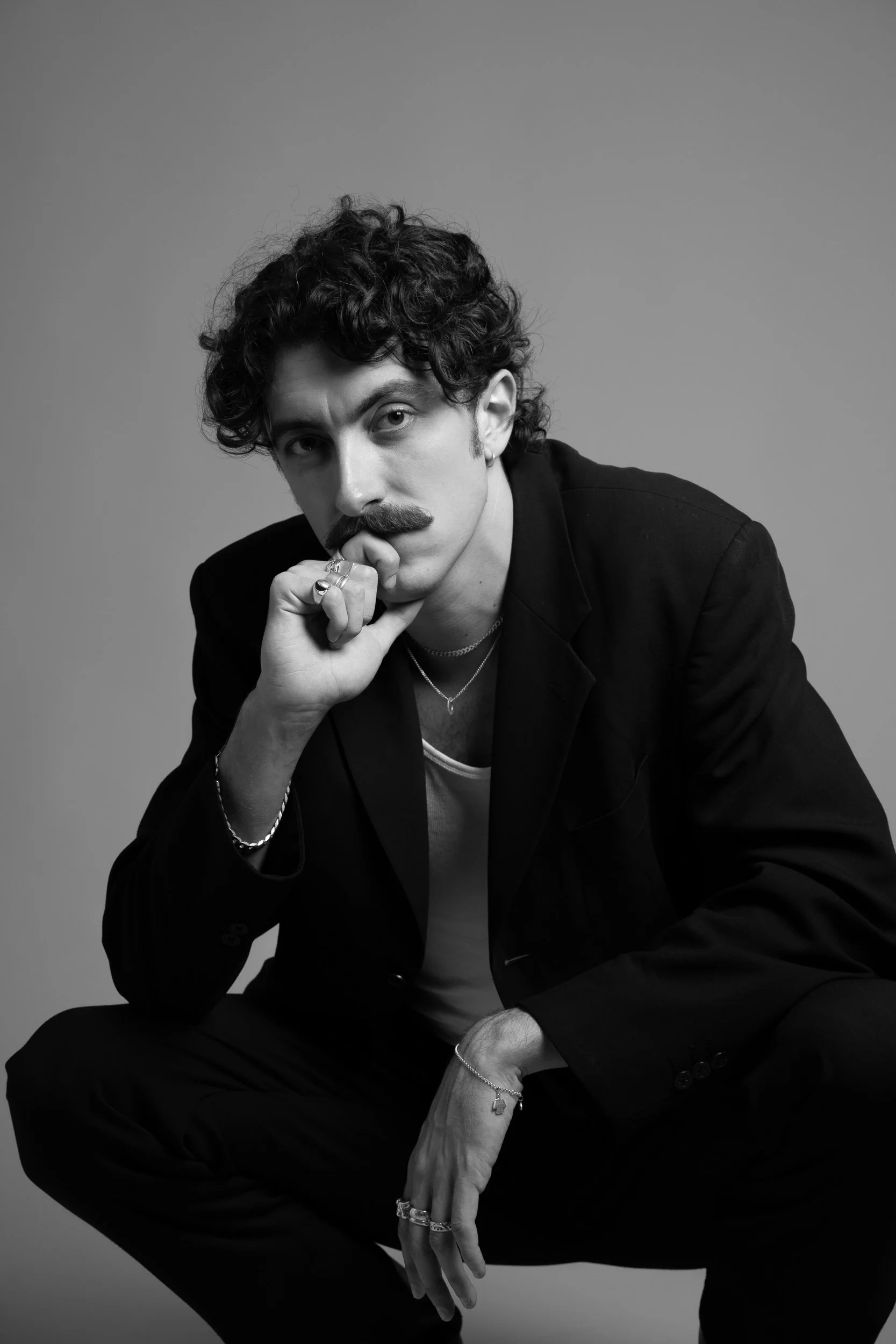 Black and white portrait of a man with curly hair, mustache, wearing rings and necklaces, dressed in a black suit, crouching with one hand on his chin and the other resting on his knee.