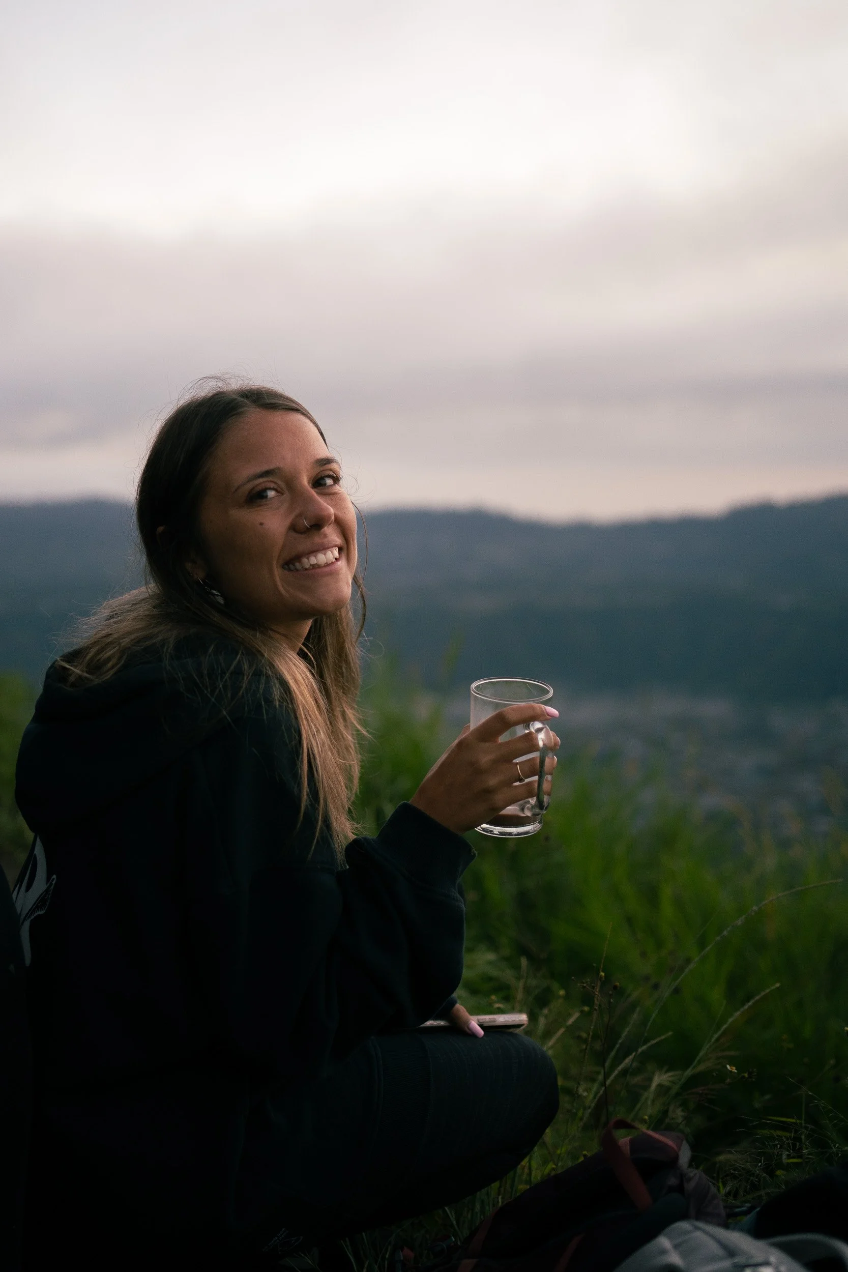 A woman with long hair, smiling, holding a glass mug, sitting outdoors in a natural setting with a cloudy sky and mountains in the background.