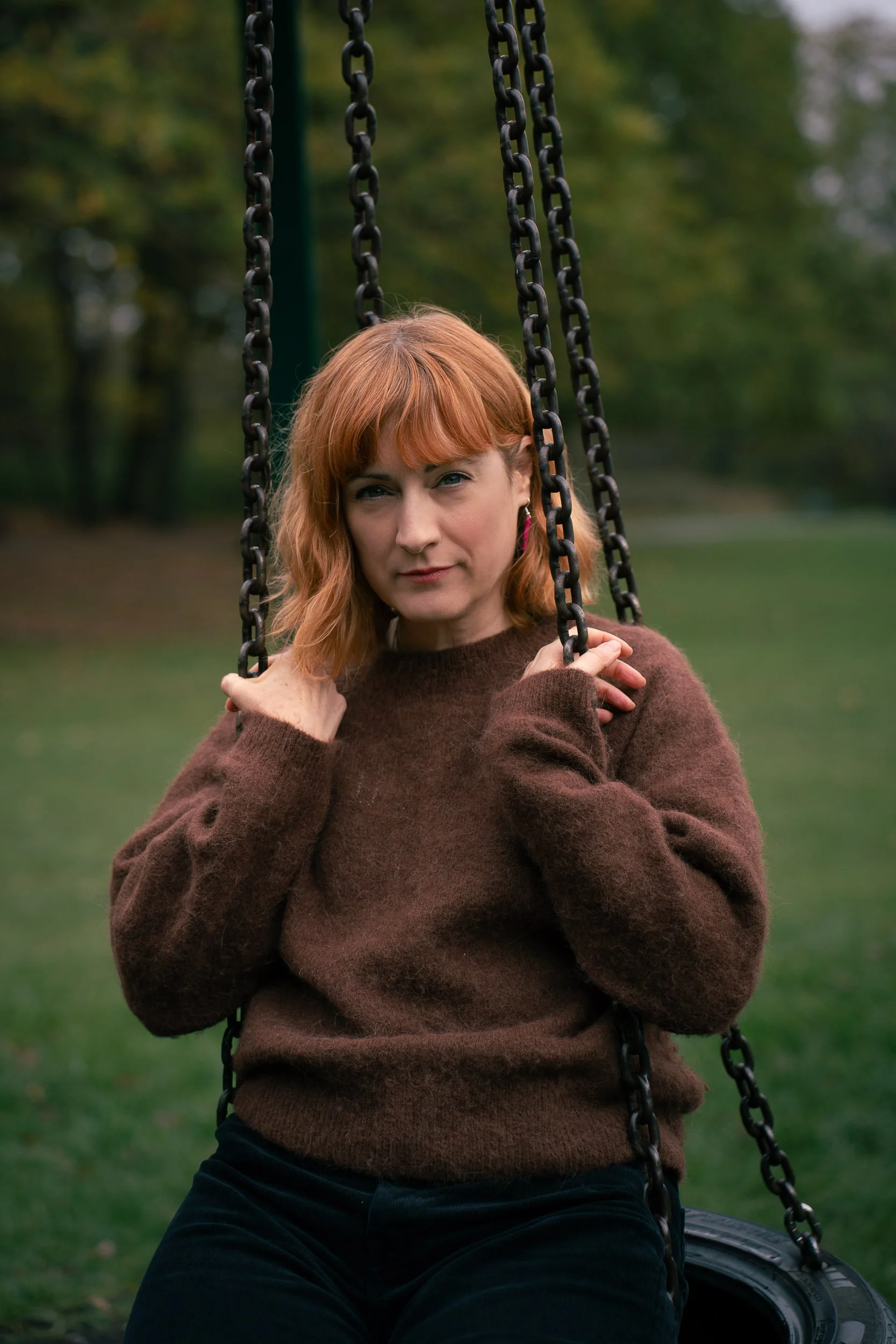 A woman with red hair and wearing a brown sweater, sitting on a swing in a park with trees in the background.