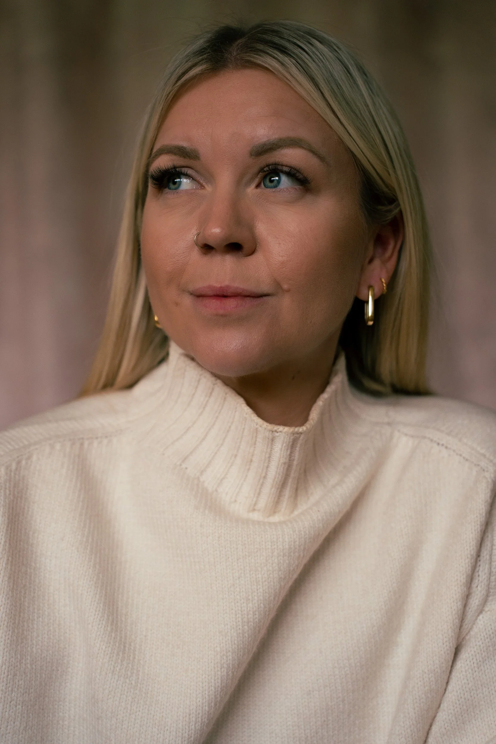 A close-up of a blonde woman with blue eyes wearing gold hoop earrings, a silver nose ring, and a cream-colored turtleneck sweater.