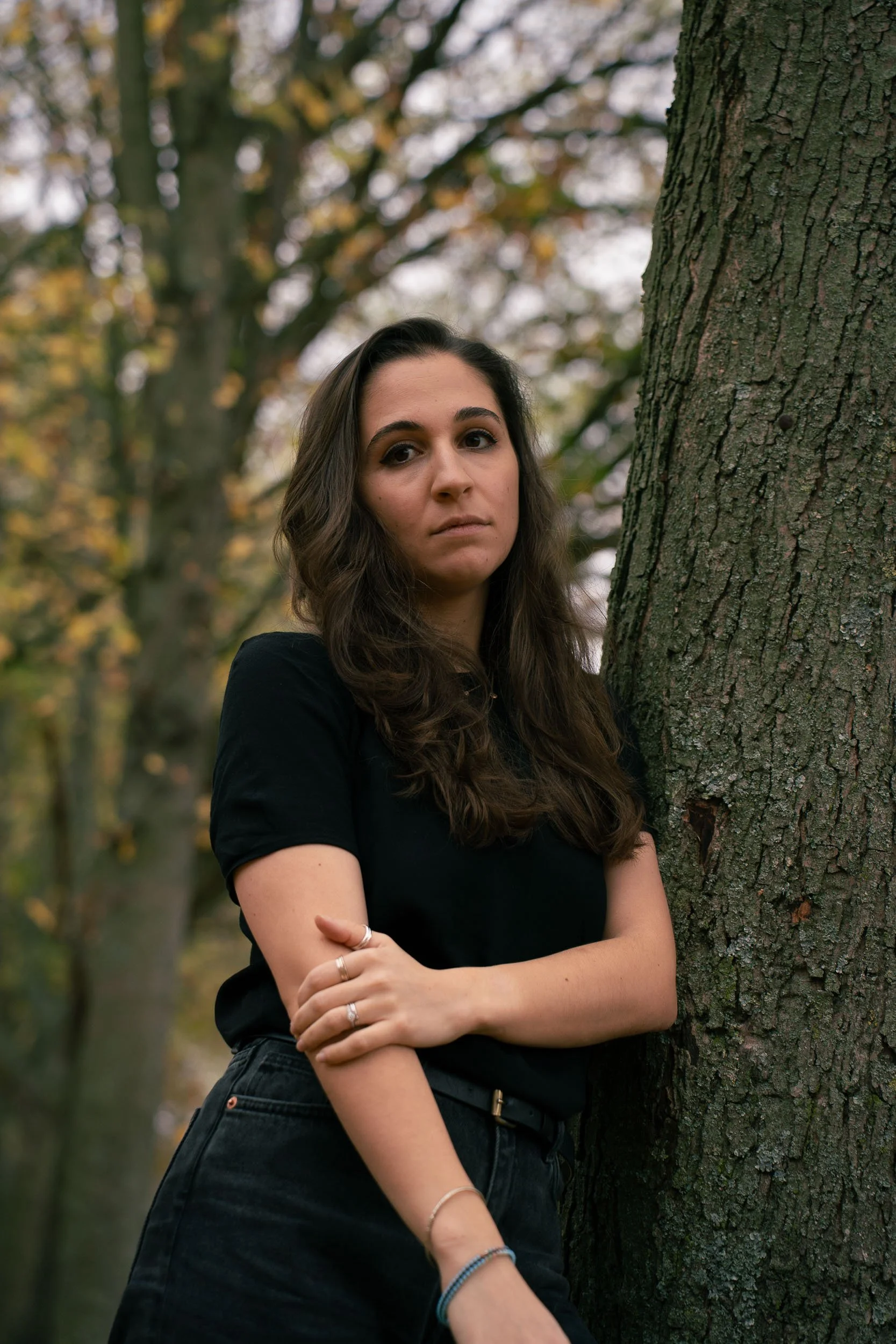 A young woman with long brown hair, wearing a black shirt and black jeans, leaning against a tree in a park with autumn foliage in the background.