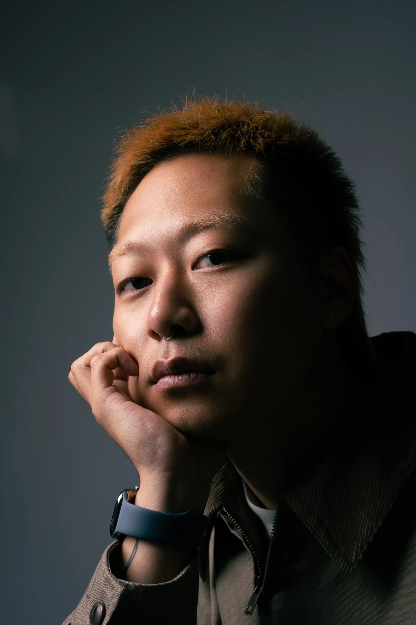 Portrait of a person with short, orange-tinted hair, wearing a septum piercing, and a smartwatch, resting their head on their hand against a dark background.
