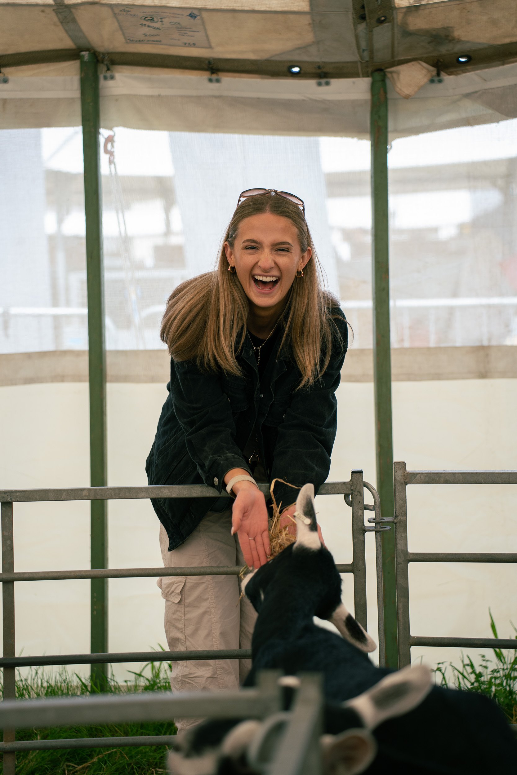 A young woman with long blonde hair, wearing glasses on her head, a black shirt, and beige cargo pants, cheerfully interacting with a black and white goat inside a barn or animal enclosure.