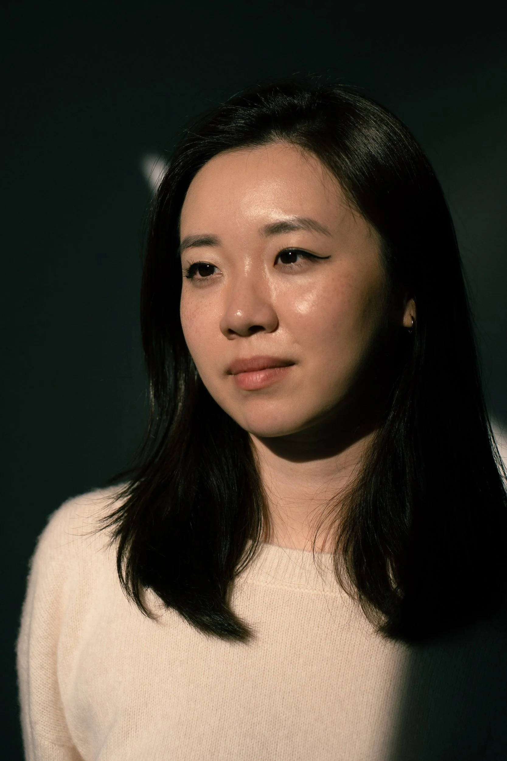 A young Asian woman with straight black hair, wearing a cream-colored sweater, looking slightly to her right with a neutral expression against a dark background.
