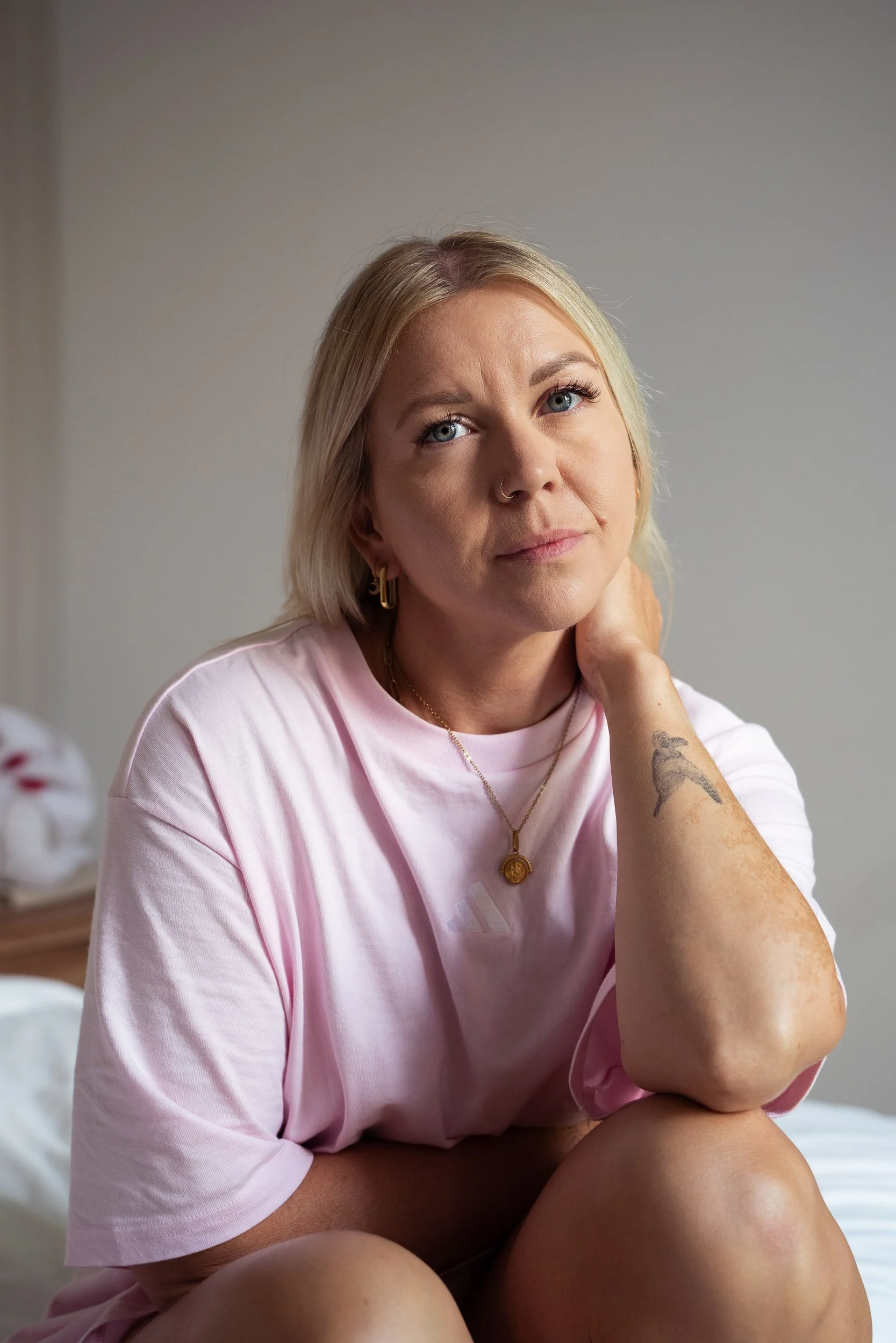 A woman with blonde hair, wearing a pink t-shirt, sitting with her knee up, looking thoughtfully at the camera, resting her chin on her hand, with a tattoo of a bird on her arm, in a softly lit room.