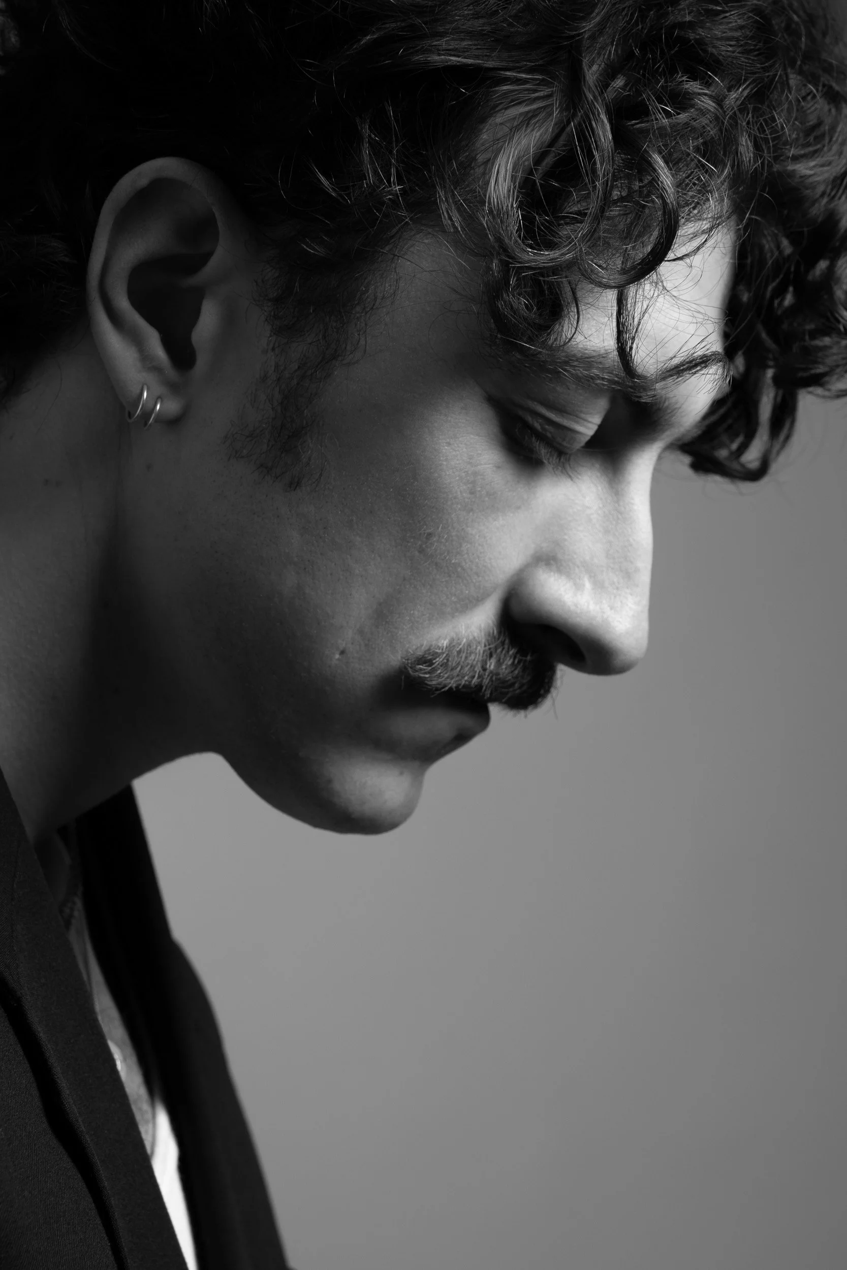 Black and white close-up portrait of a man with curly hair, mustache, and earrings, in profile, with eyes closed.