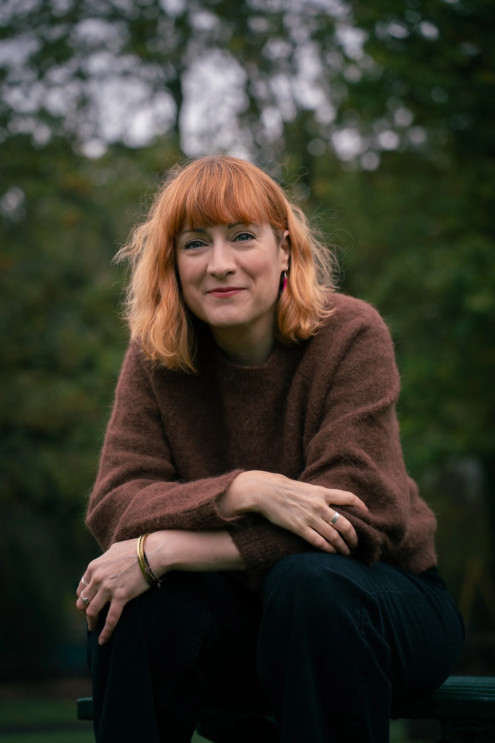 A woman with red hair sitting outdoors in a park, smiling at the camera, wearing a brown sweater and black pants.