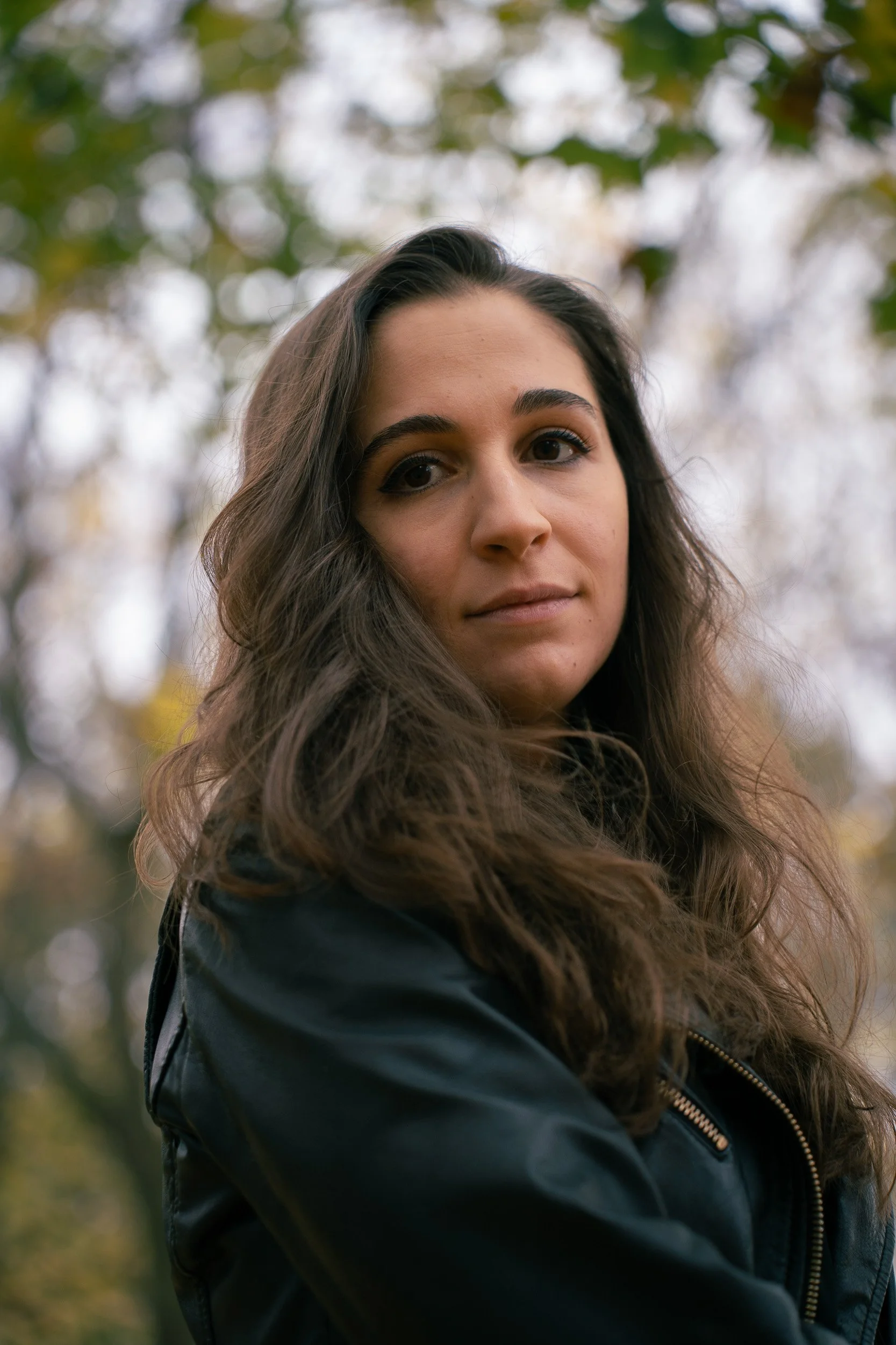 A young woman with long wavy brown hair and light skin looking into the camera outdoors, wearing a black jacket with a zip, with blurred autumn trees in the background.
