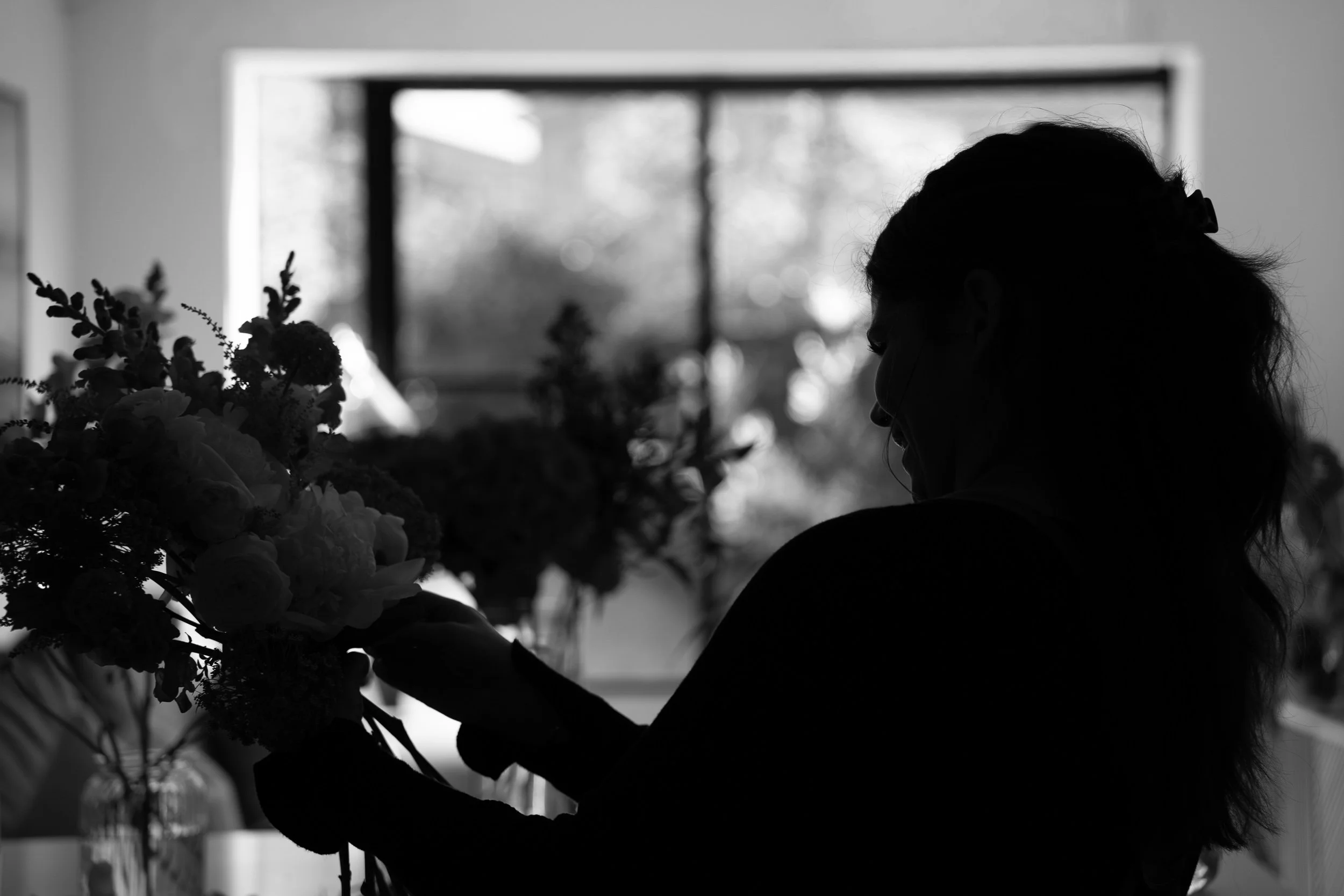 Silhouette of a woman arranging flowers near a window.