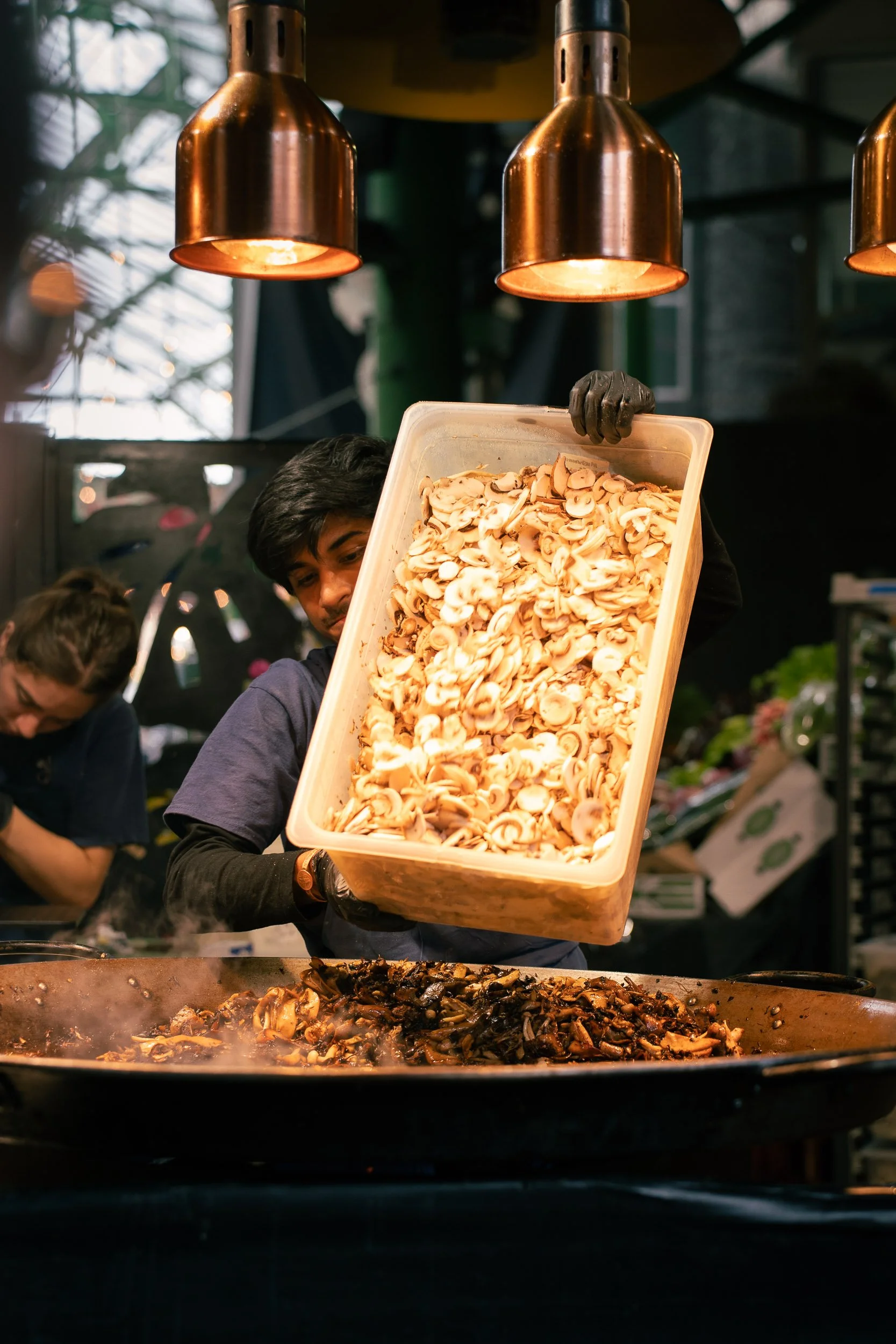 A person wearing a black glove holds a large container of sliced mushrooms over a cooking pan with mushrooms, with a woman in the background in a busy kitchen or food stall under warm lighting.