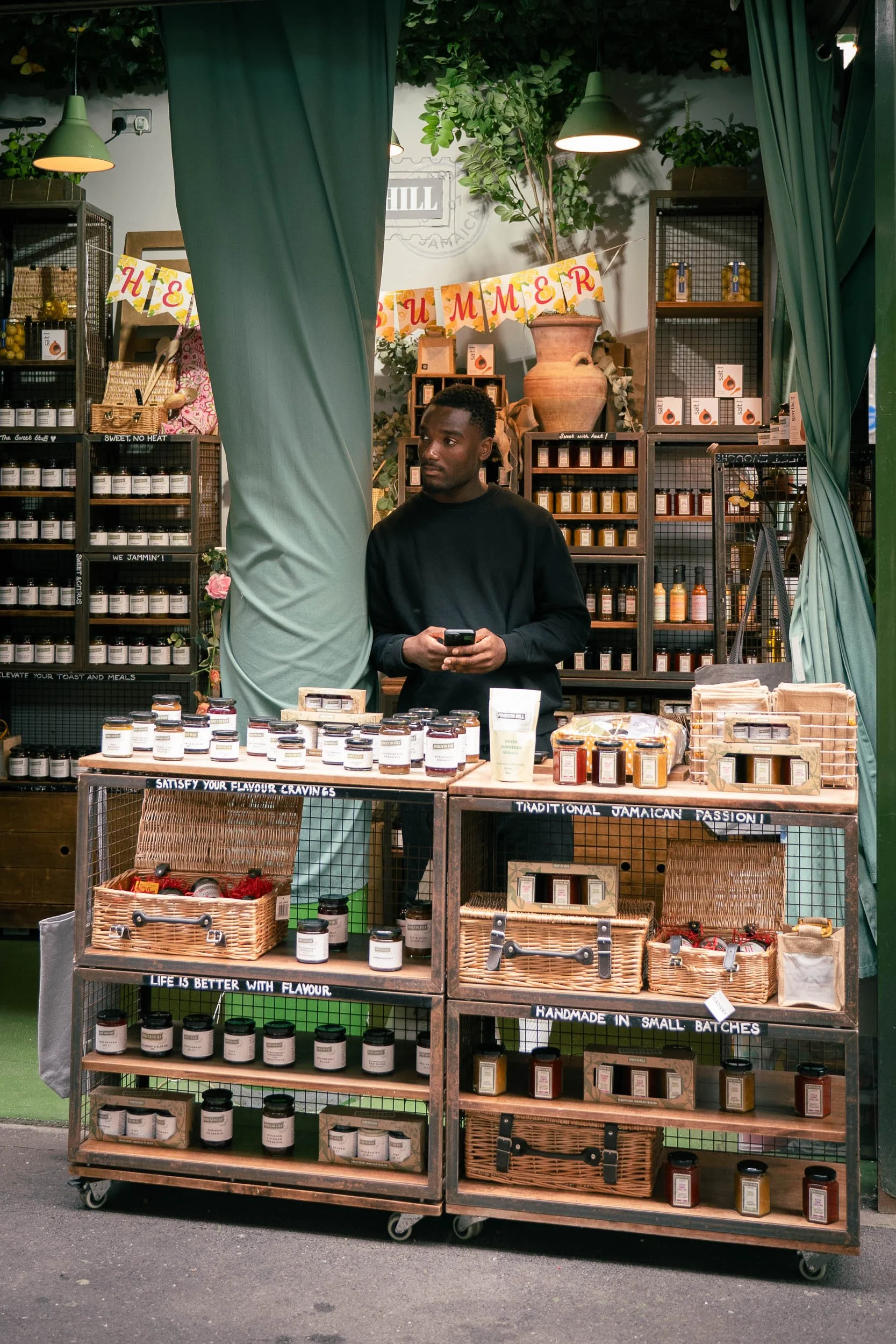 A man standing behind a stall at an outdoor market, selling jars of flavored products with a tropical theme, decorated with green curtains, shelves stocked with jars, and a "Summer" banner.