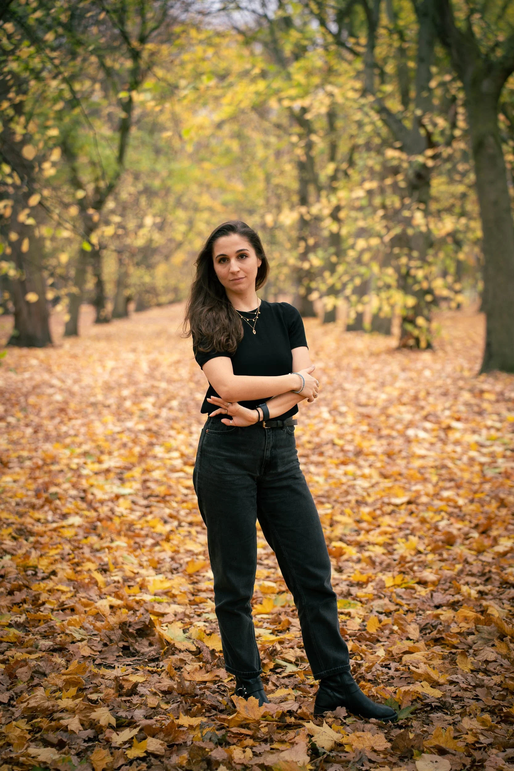 A woman standing in a forest with fall foliage, wearing a black shirt, black jeans, and black boots.