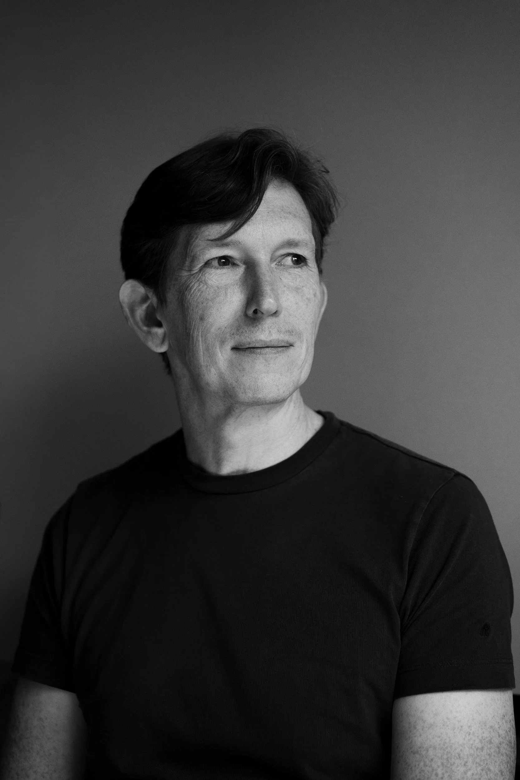 Black and white portrait of a man with short hair, wearing a dark t-shirt, looking off to the side against a plain background.