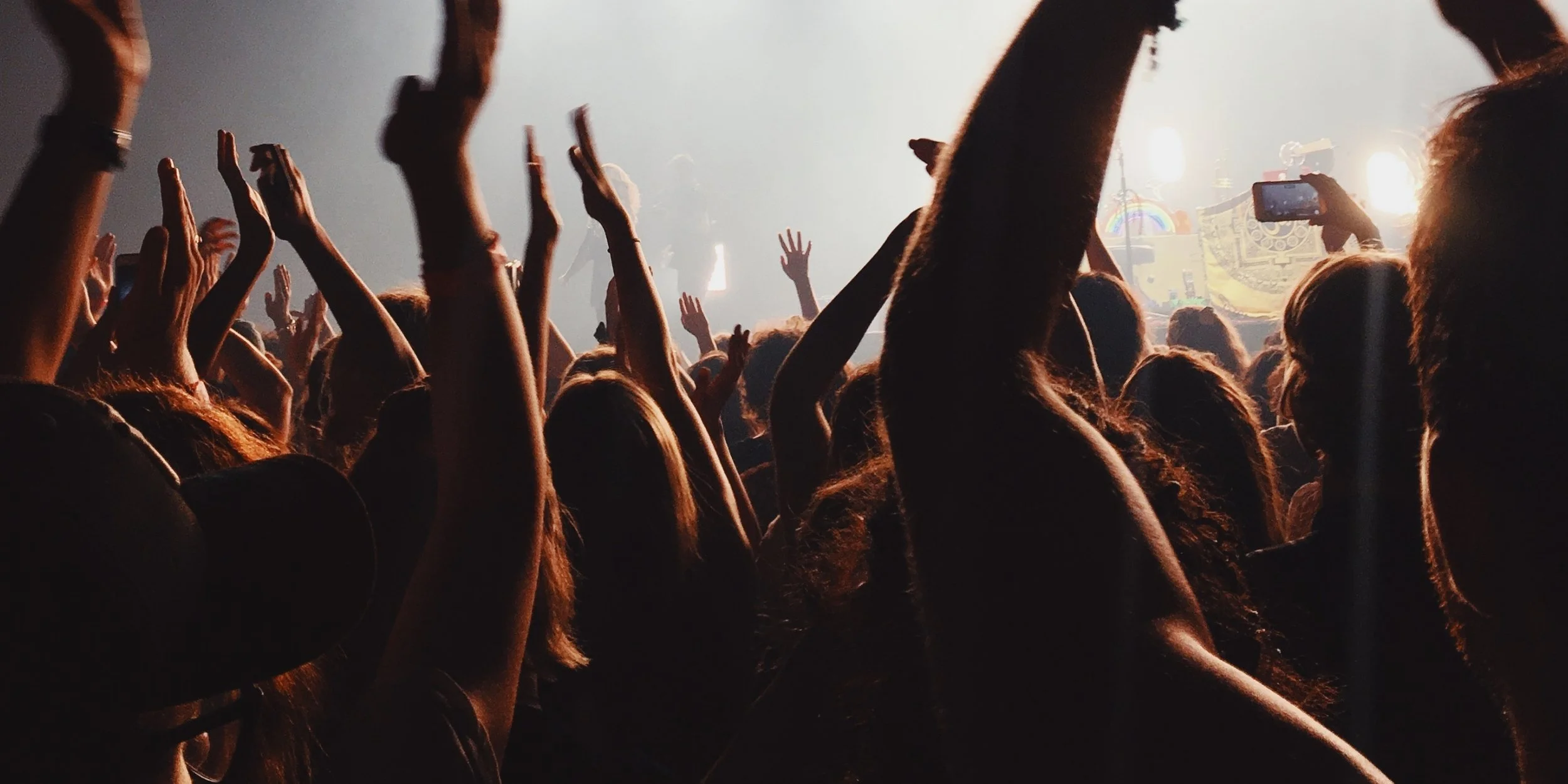 Crowd at a concert with hands raised, stage lights in the background, and some people taking photos with their phones.