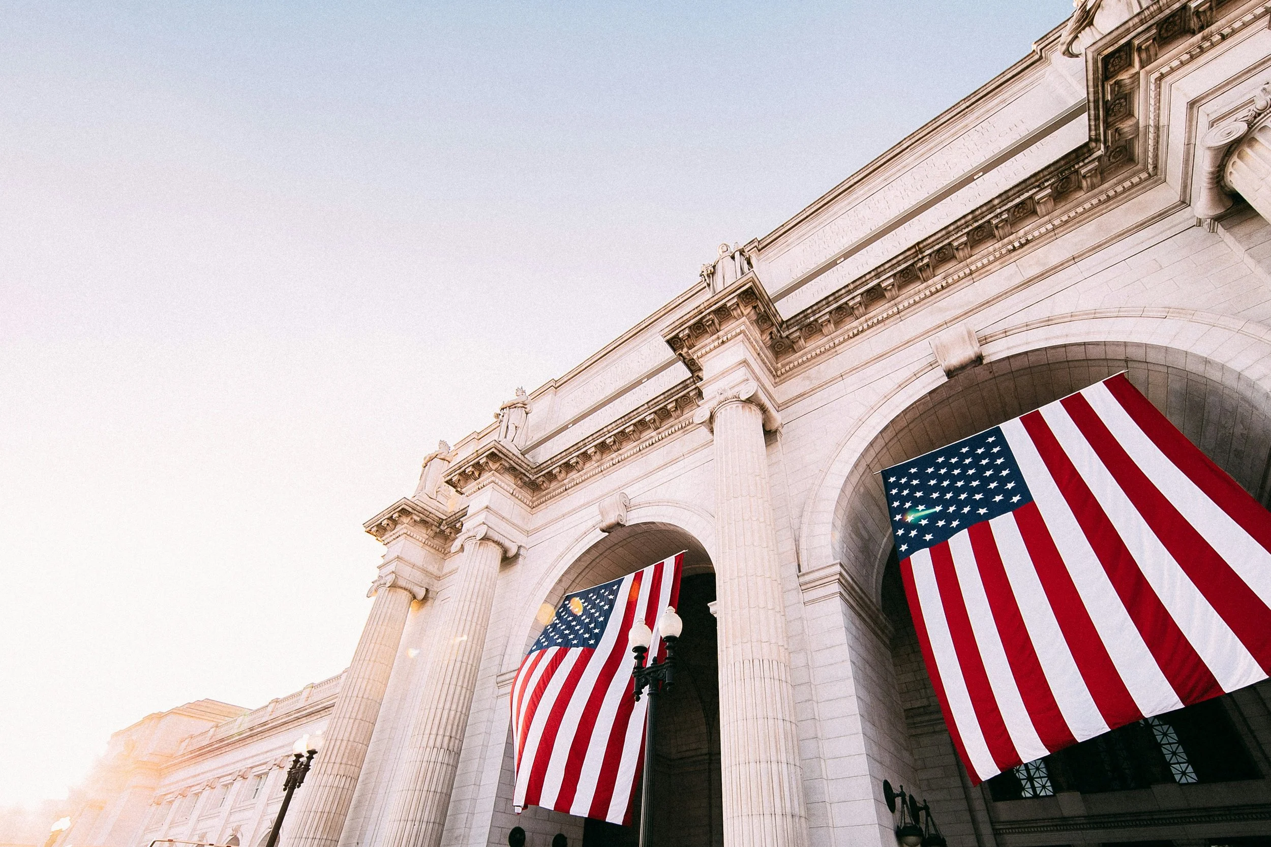 The image shows an upward view of a historic neoclassical building with large columns, decorated with American flags hanging from the archways.