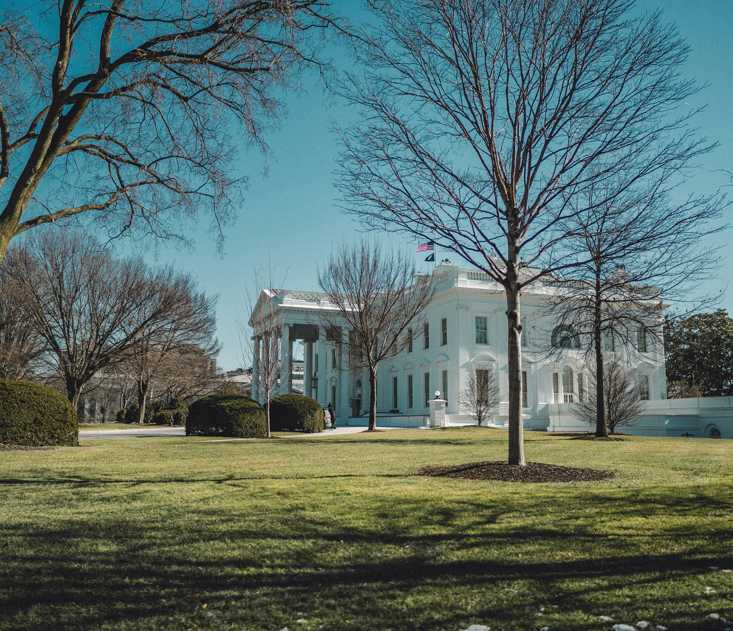 The White House, the official residence of the U.S. president, viewed from the front lawn during daytime. The scene includes leafless trees, well-maintained grass, and clear blue skies.