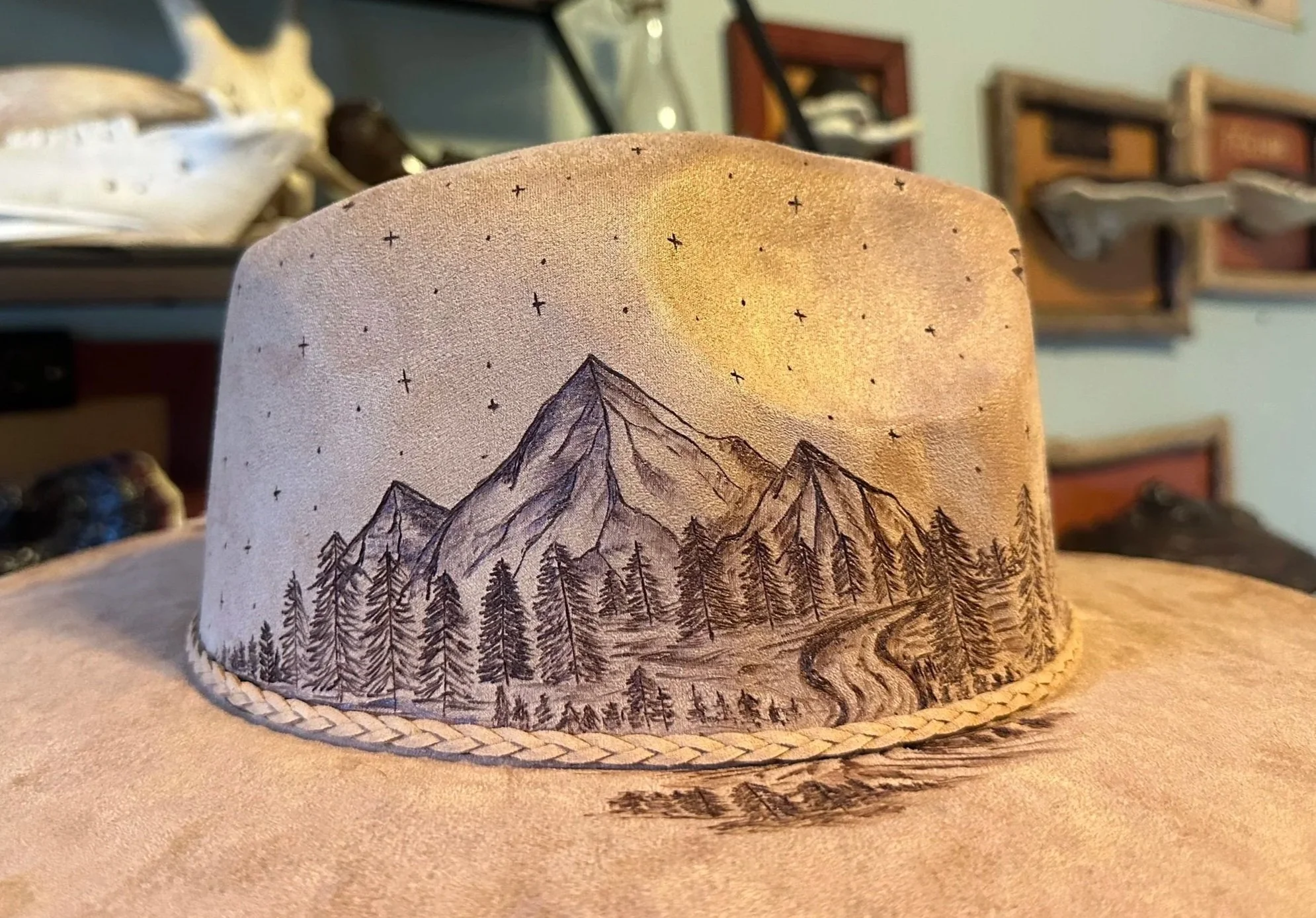 A beige vegan suede rancher fedora with a mountain and forest landscape drawing