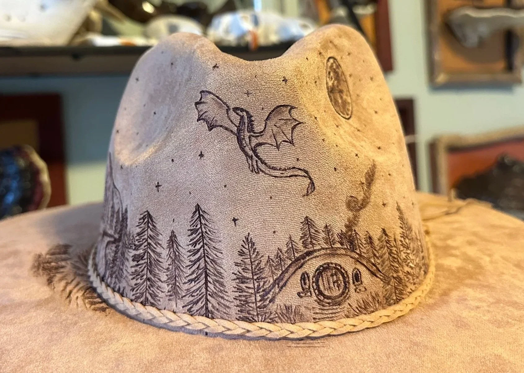 A vegan suede rancher fedora hat with a hand-burned dragon, forest, full moon and hobbit house.