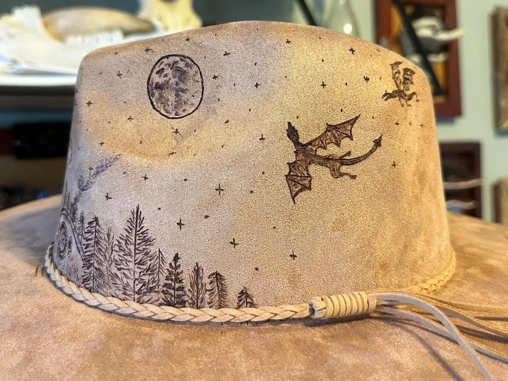 A vegan suede fedora hat with a moon, stars, trees, and two flying dragons hand burned on.