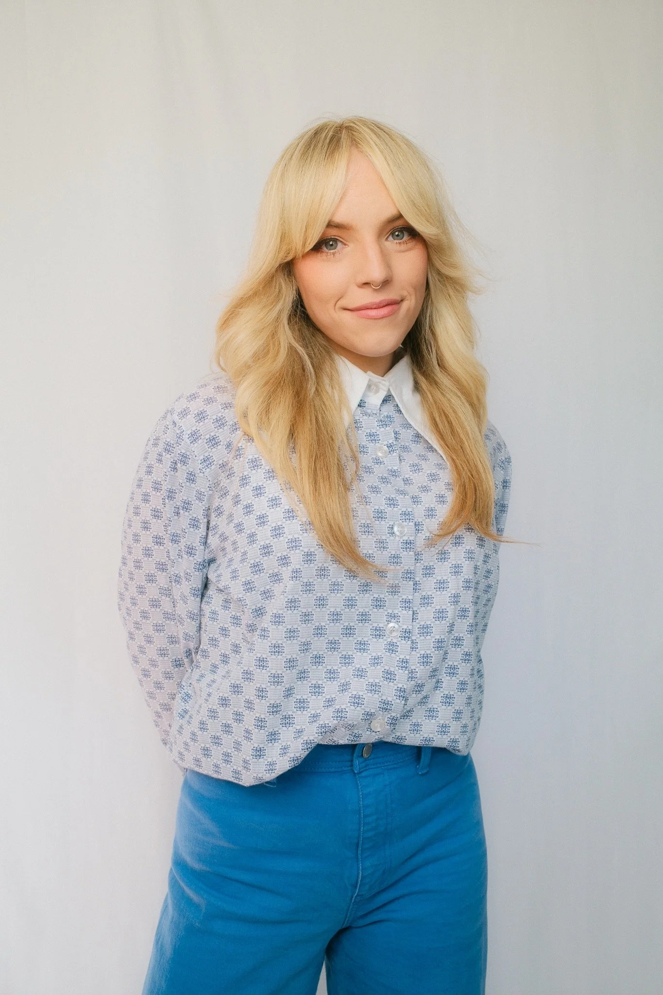Young woman with blonde wavy hair, wearing a light-colored patterned blouse with a white collar and blue high-waisted pants, smiling at the camera.