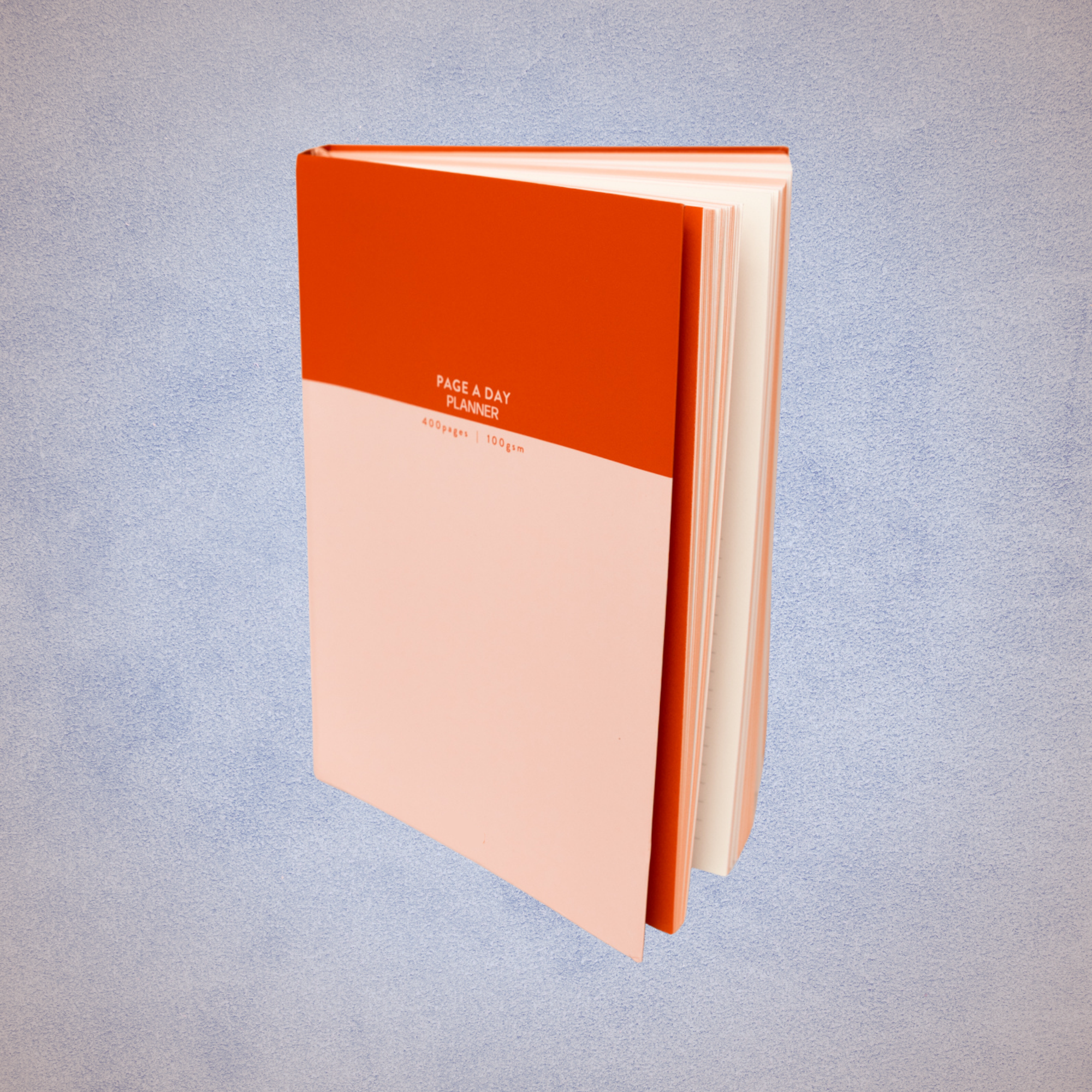 Open planner notebook with orange and light pink cover, titled 'Page a Day Planner' with 400 pages and 100 gsm paper, standing upright on a textured light blue background.