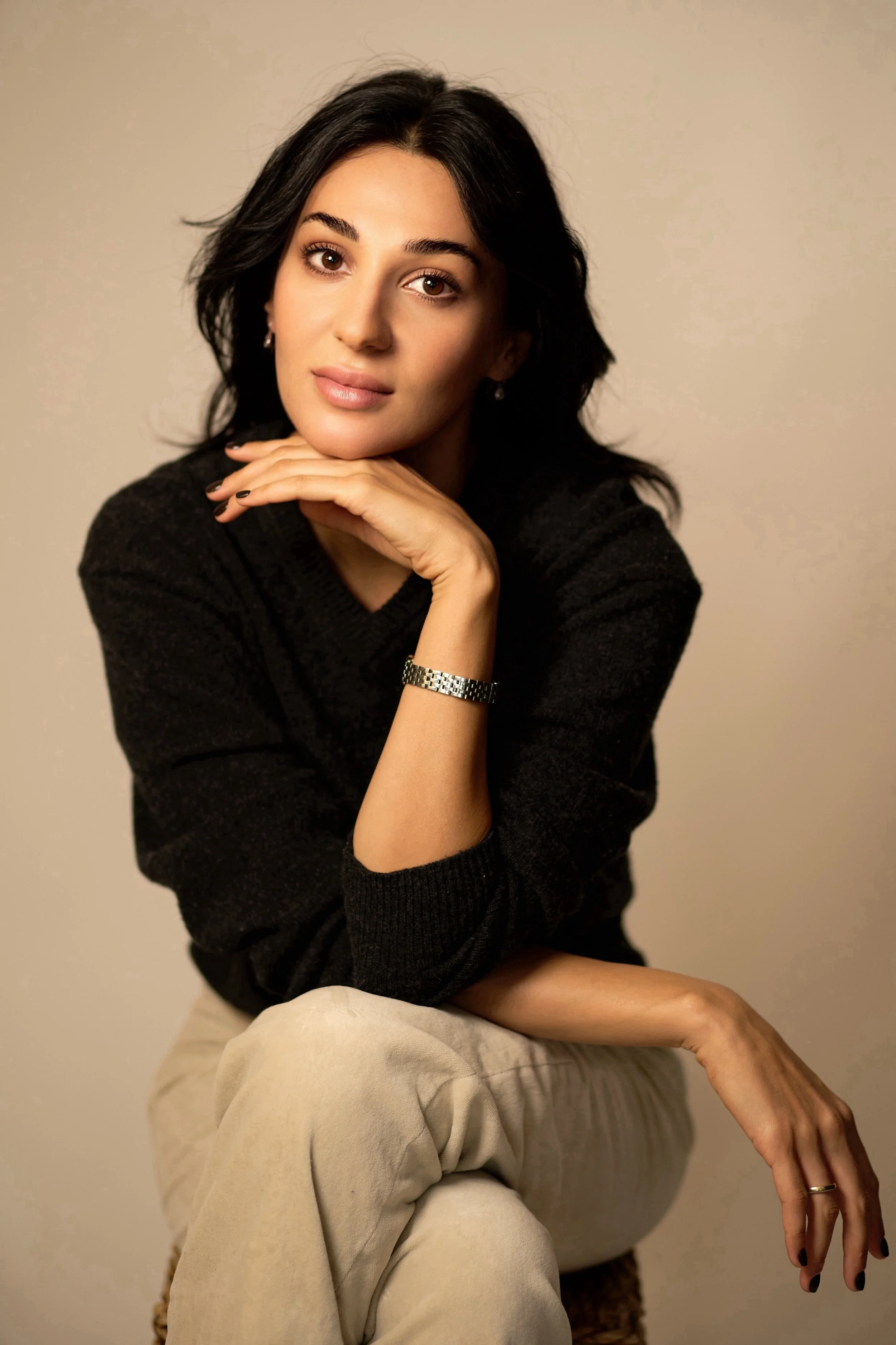 Portrait of a young woman with black hair, wearing a black sweater, beige pants, and jewelry, sitting against a neutral background.