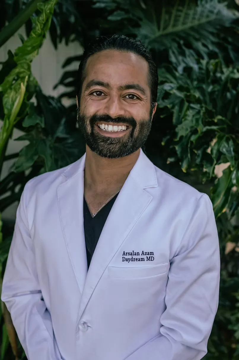 A smiling man with dark hair and a beard wearing a white  medical coat with the text "Arsalan Azam Daydream MD" on it, standing outdoors in front of green leafy plants.