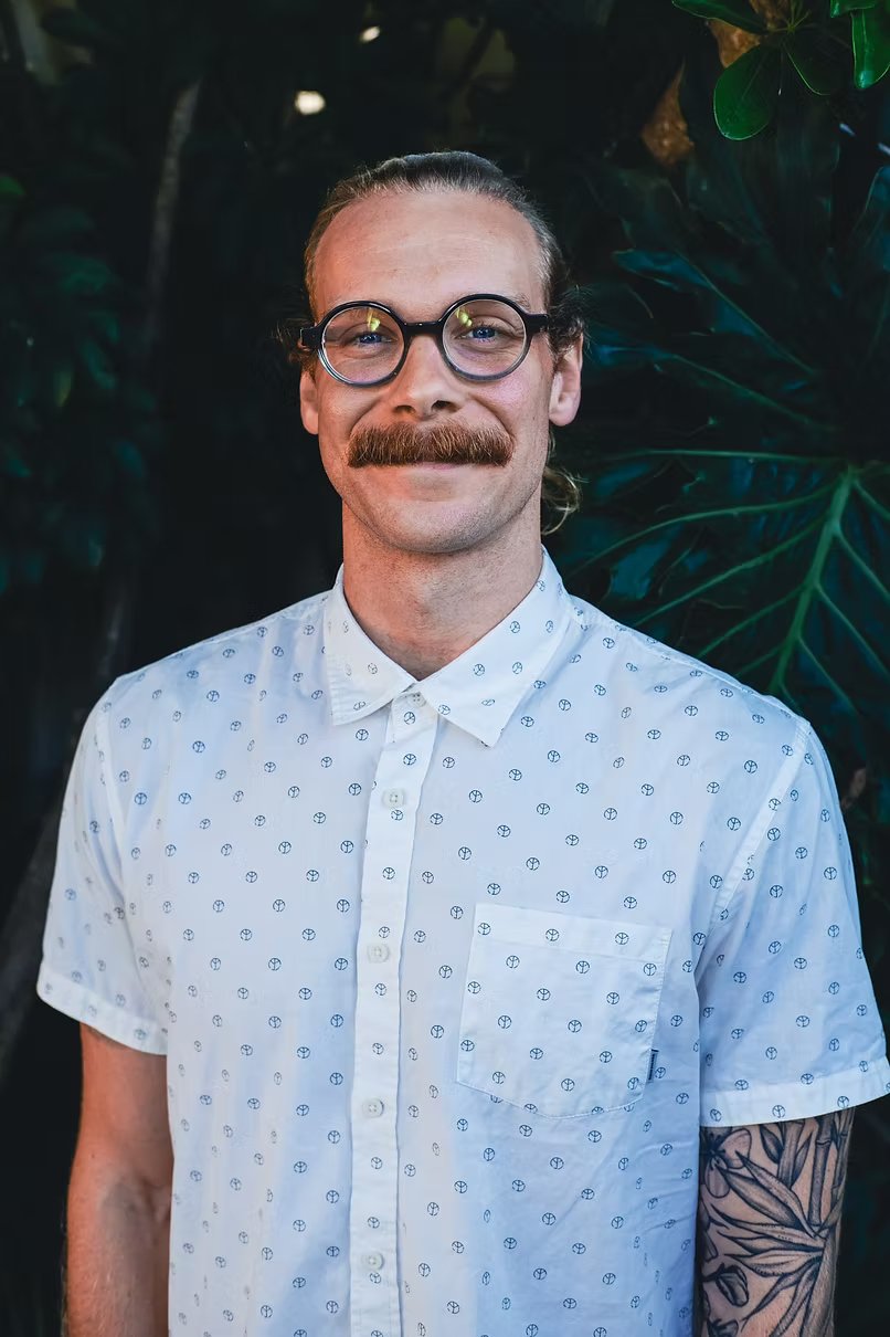 A man with glasses and a mustache, smiling, wearing a white shirt with small blue symbols, standing outdoors with green foliage in the background.