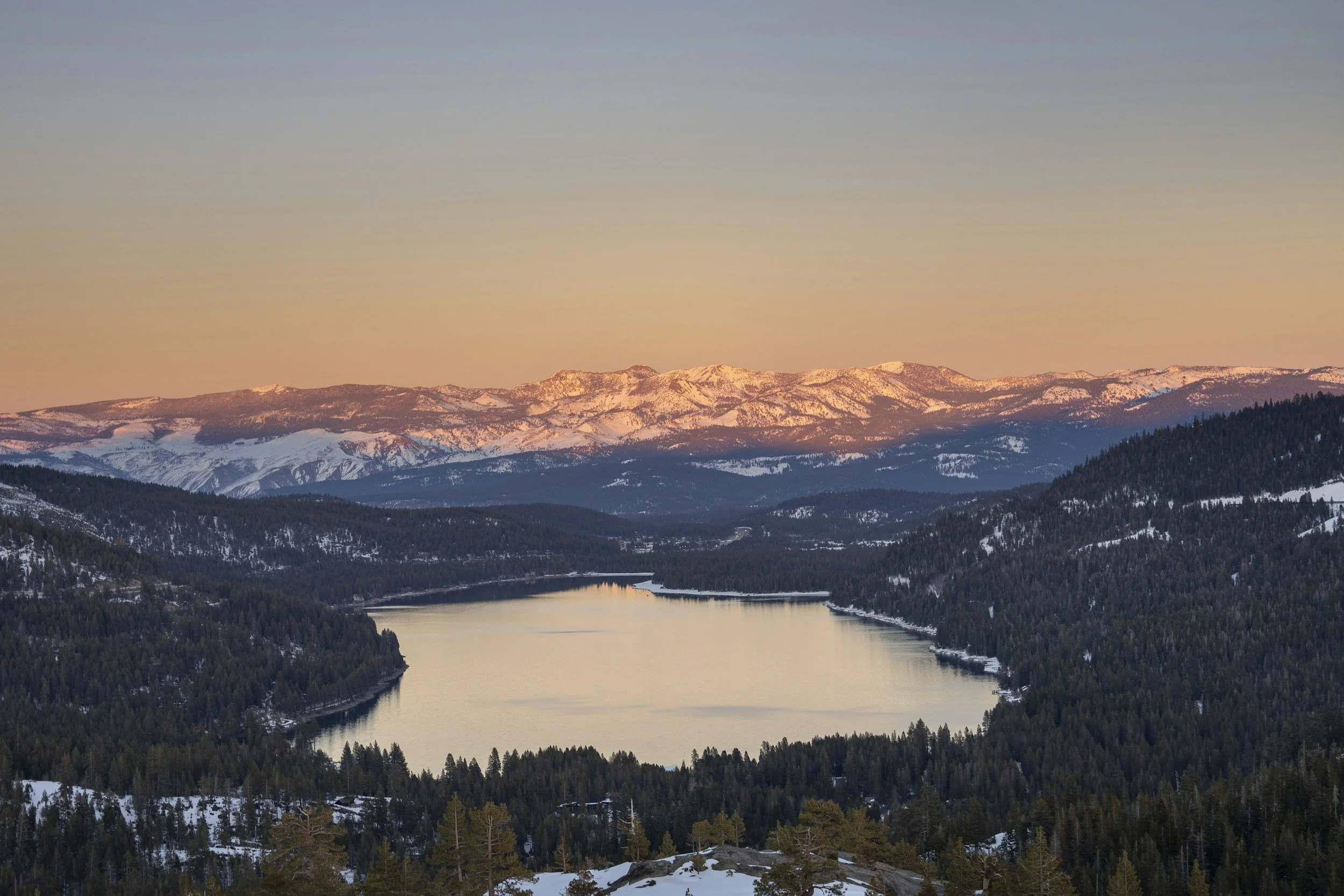Sunset over snow-capped mountains with a river in a forested valley.