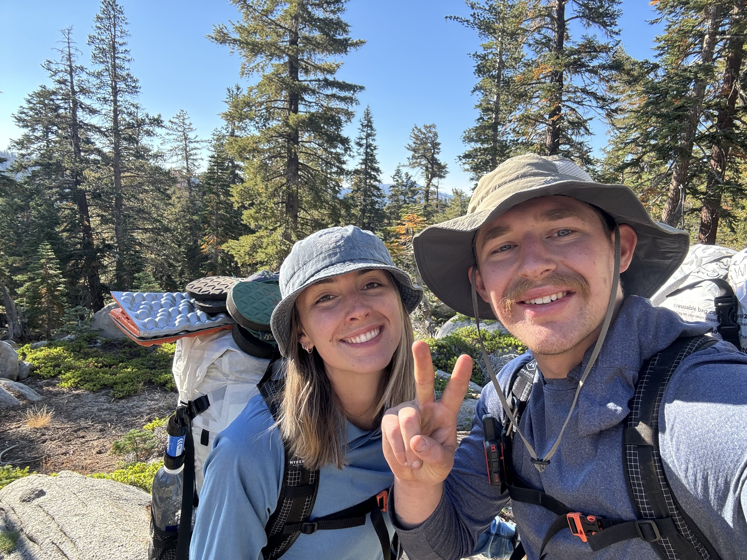 Two hikers smiling and making a peace sign in a scenic forest with tall pine trees on a sunny day.