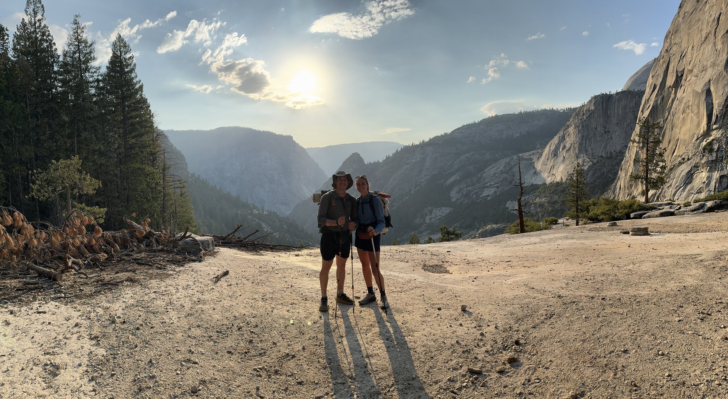 Two hikers standing on a dirt trail in a mountainous forested area with cliffs and trees, during the daytime with the sun shining in the sky.