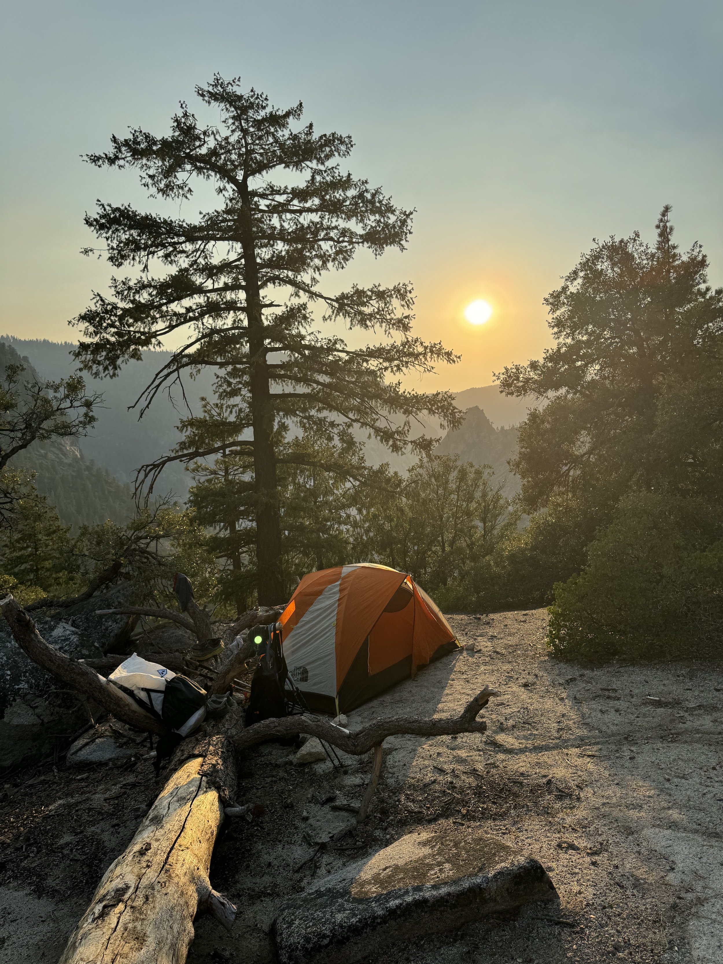 A campsite with an orange and white tent set up on a dirt patch among trees, with a fallen log and backpacks nearby, during sunset in a forested mountainous area.