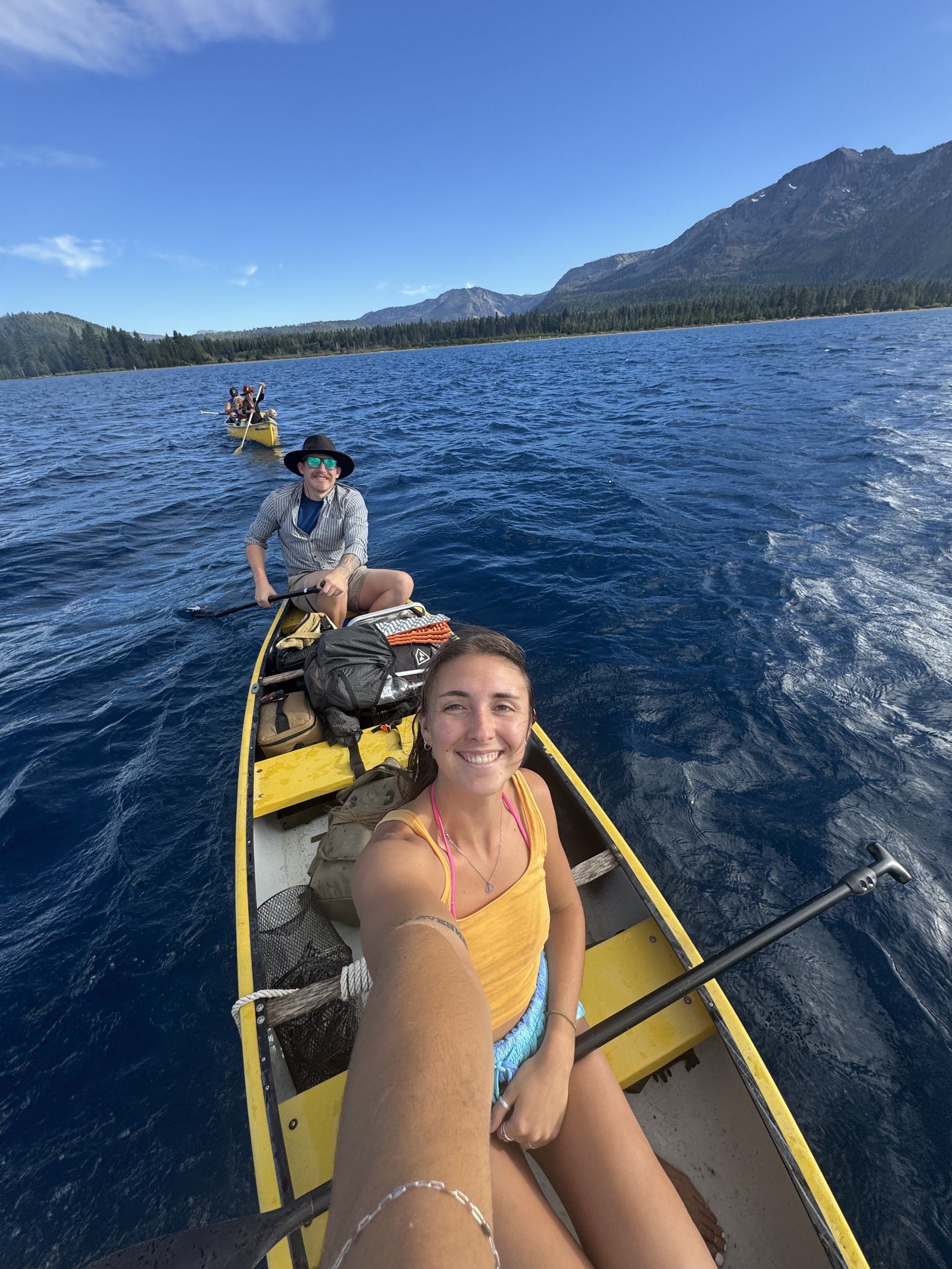 Three people on a yellow canoe in a mountain lake, with forested hills and mountains in the background on a clear, sunny day. The woman in the front is smiling for a selfie, wearing a yellow tank top and shorts, with a paddle in her hand. The man beh