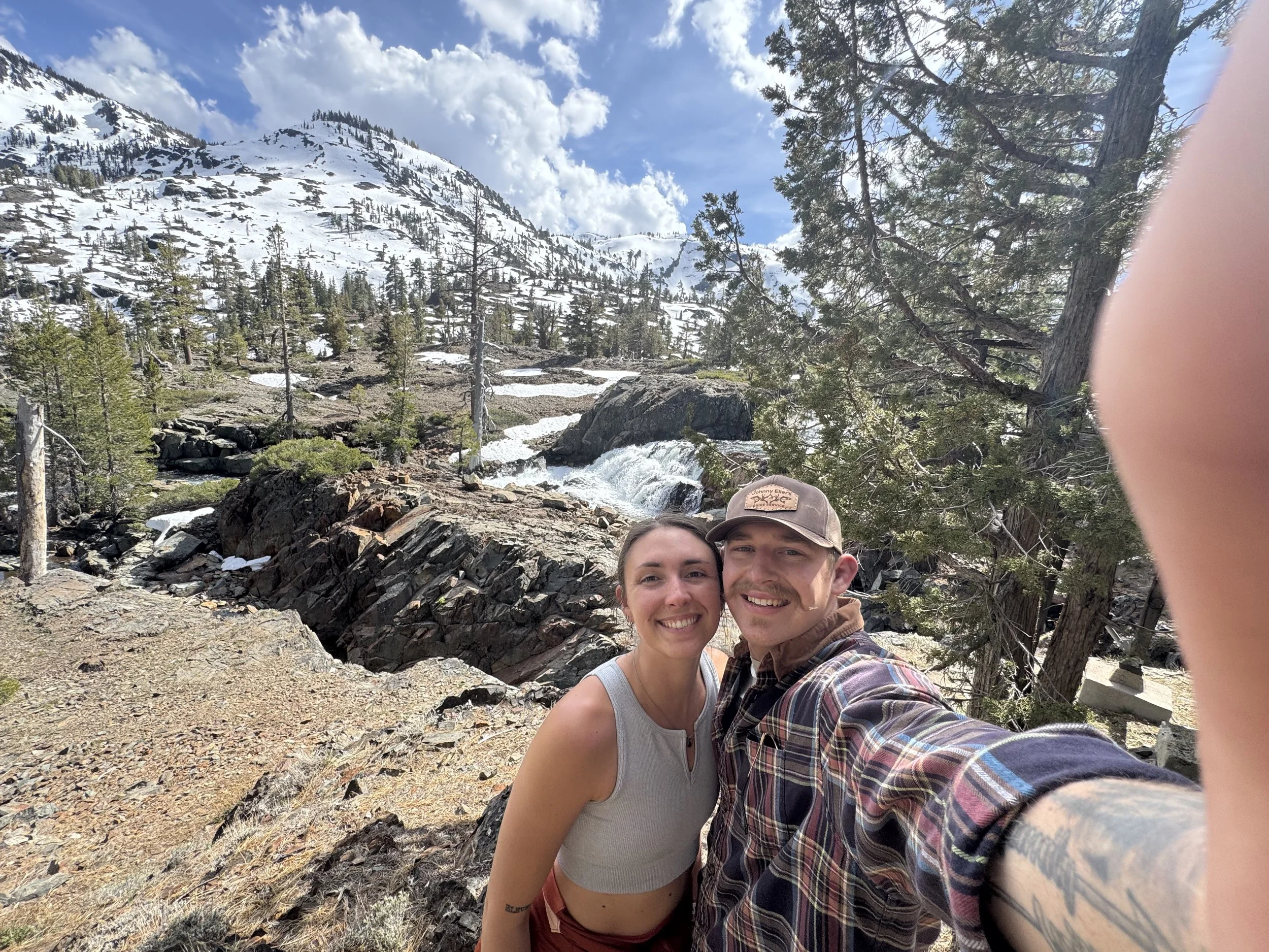 A smiling couple takes a selfie outdoors in a mountainous, forested area with snow, trees, rocks, a small waterfall, and a cloudy sky in the background.