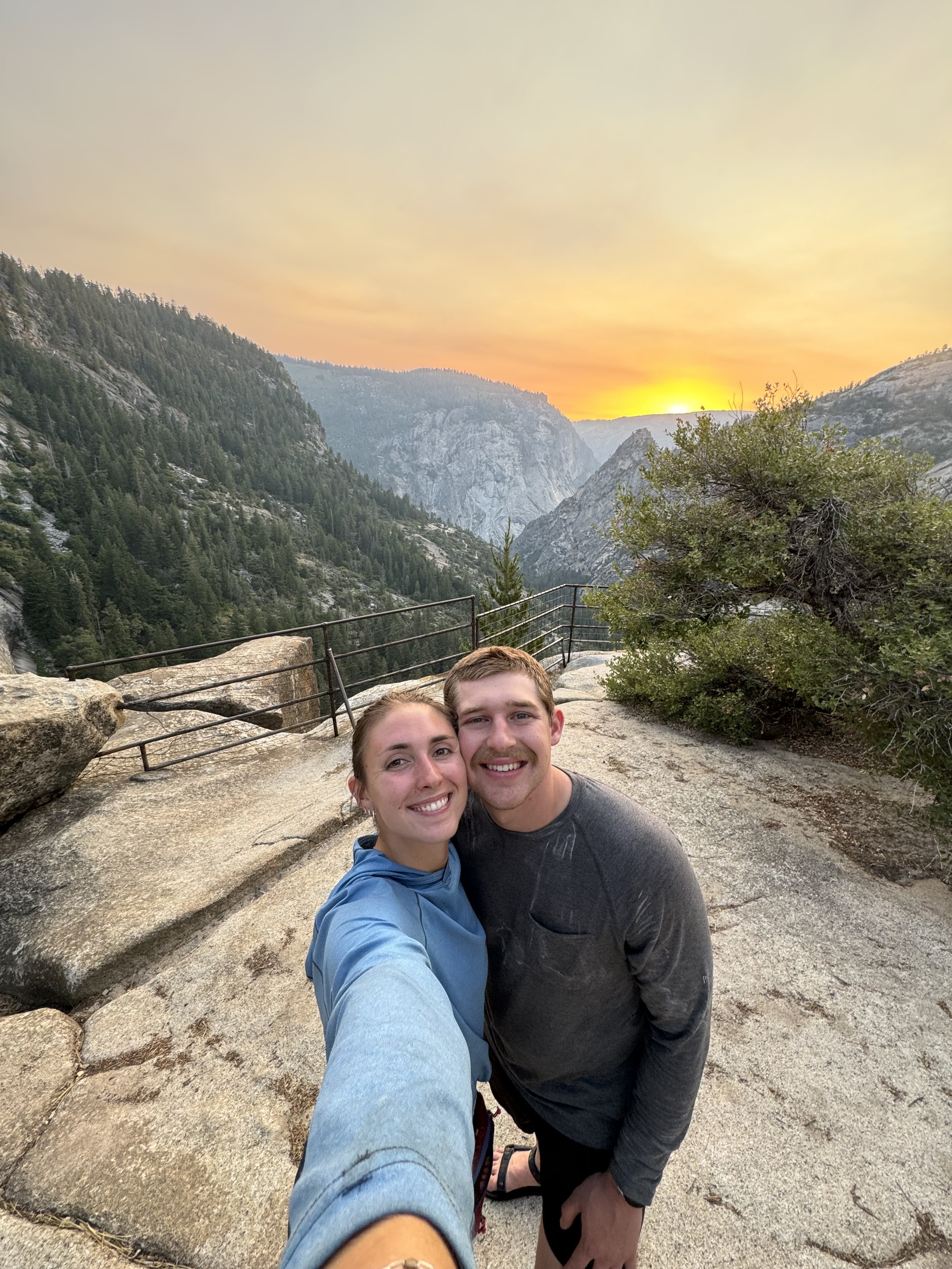 A smiling couple taking a selfie on a rocky overlook at sunset surrounded by mountainous landscape and trees.