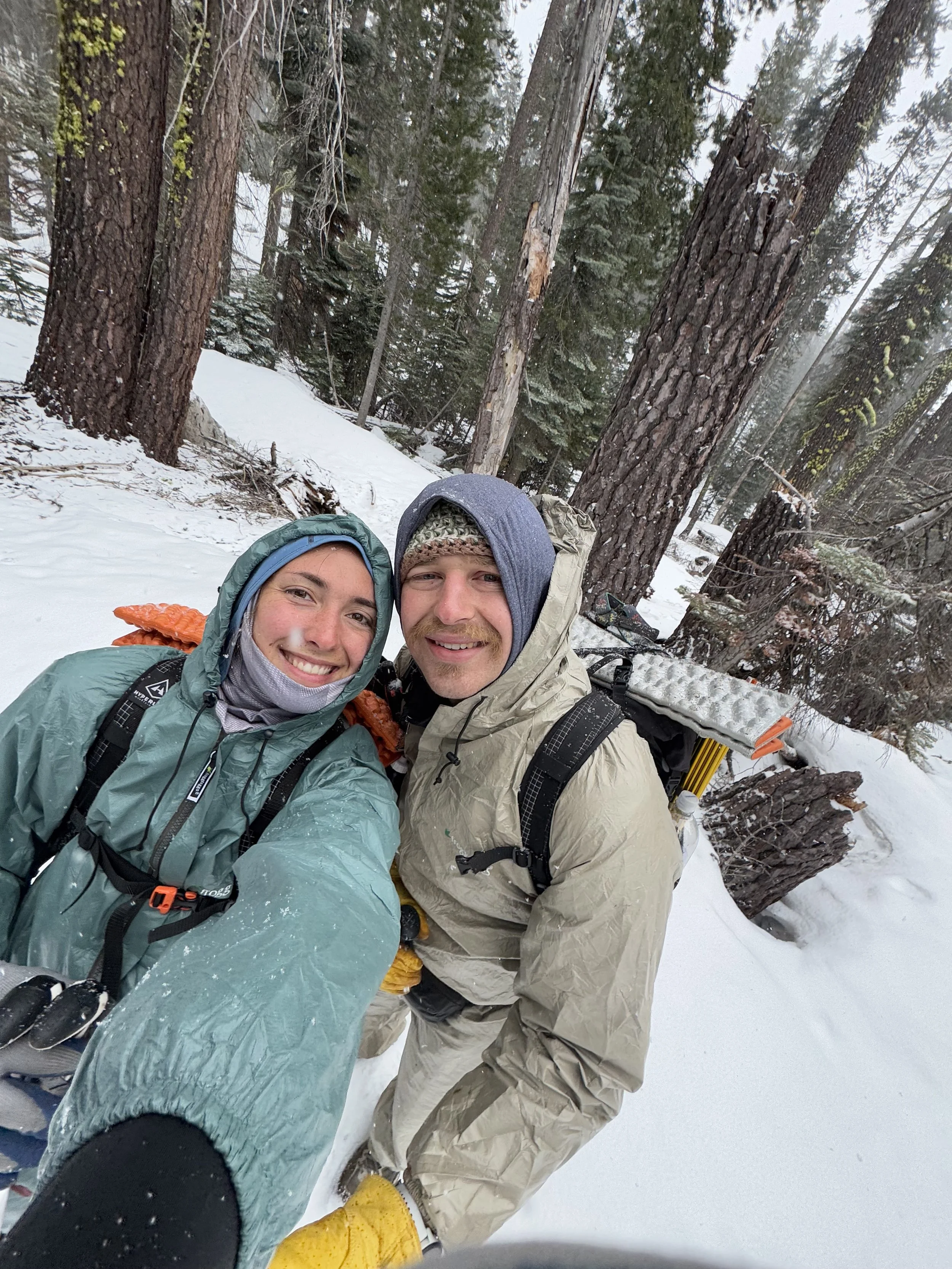 Smiling couple taking a selfie outdoors in a snow-covered forest, dressed in winter gear with trees in the background.