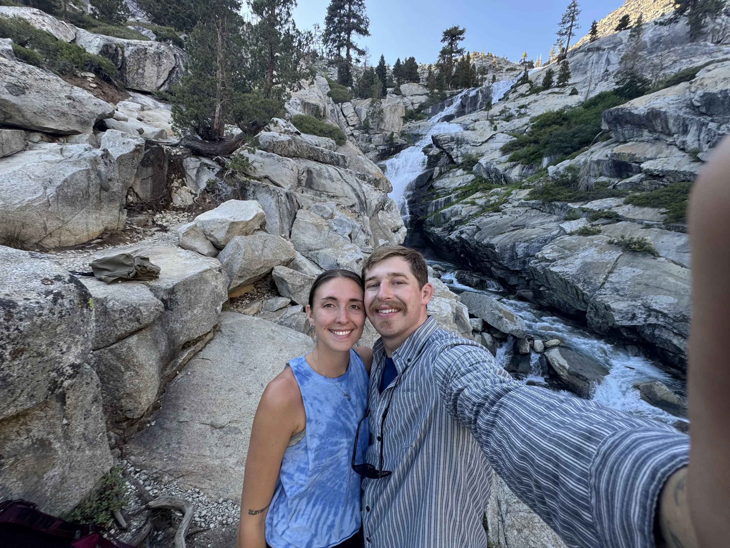 A young couple taking a selfie in front of a waterfall and rocky landscape with trees.
