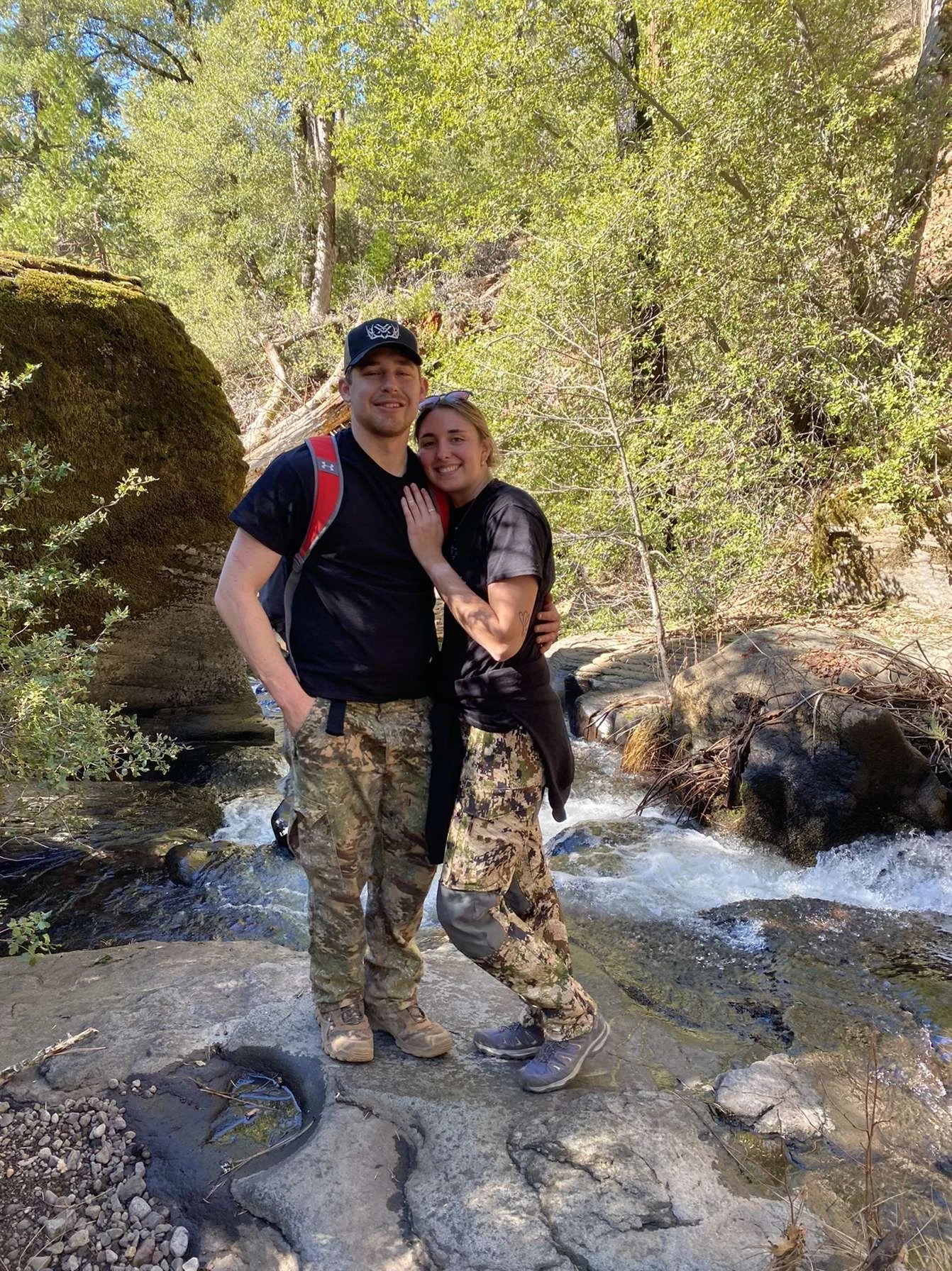A couple standing on a rock in a creek, surrounded by trees and greenery, smiling at the camera.