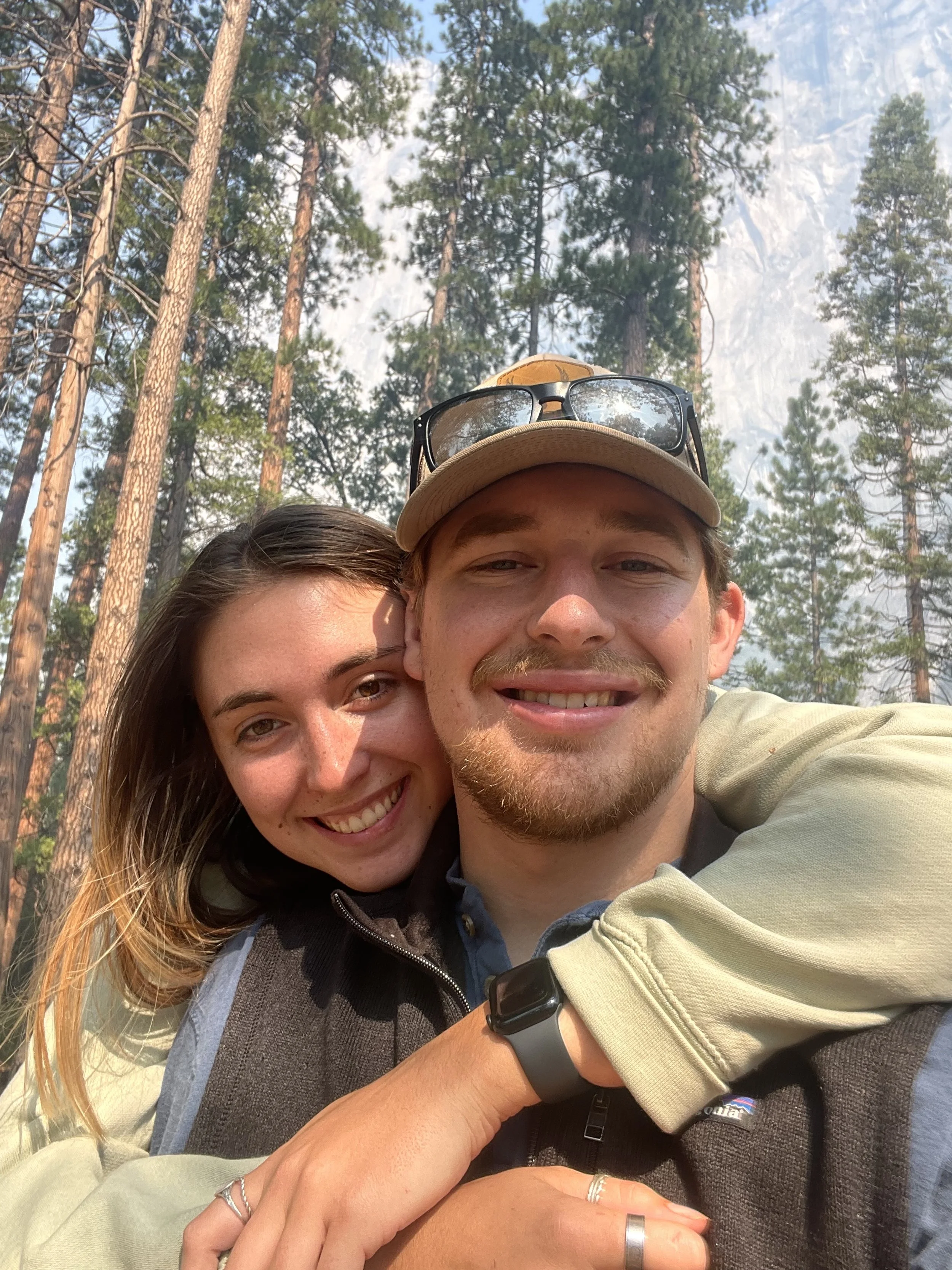 A young couple smiling and embracing outdoors with tall pine trees in the background.