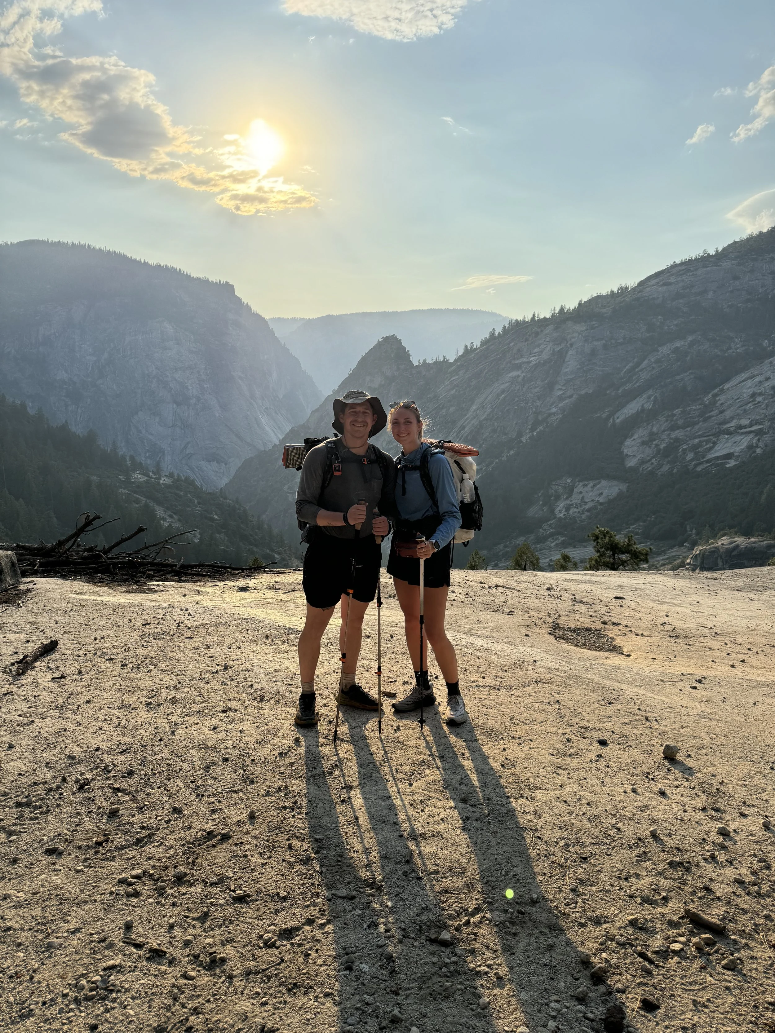 Two hikers standing together on a dirt trail in a mountainous area, with backpacks and trekking poles, under a sunny sky with scattered clouds.